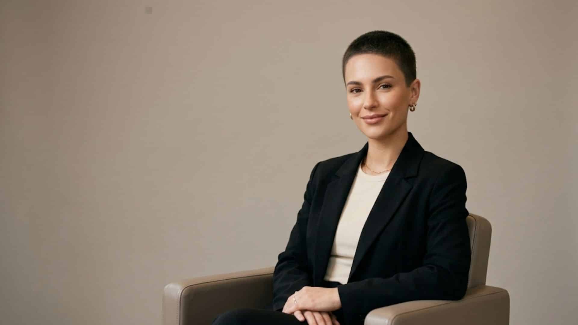 Woman with a buzzed Caesar cut hairstyle, sitting in a black blazer smiling softly