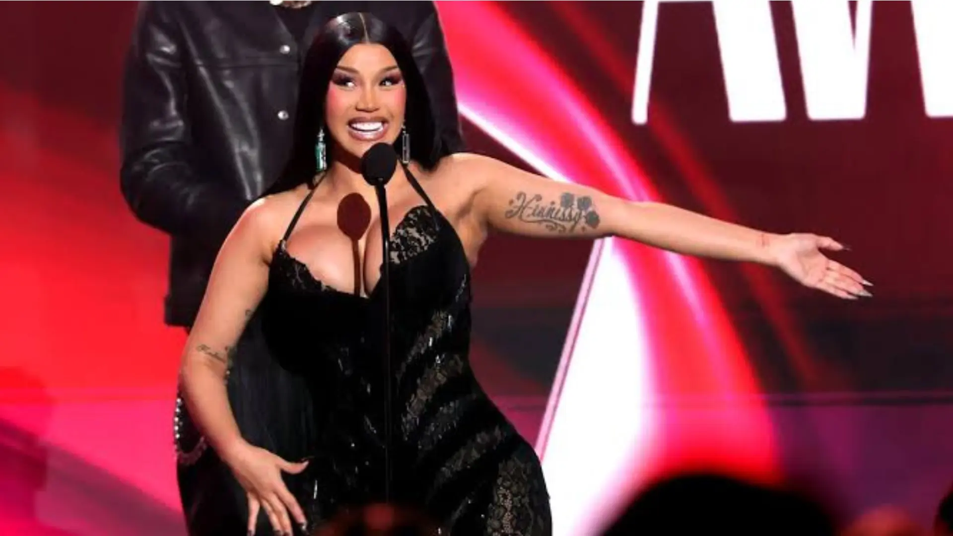Cardi B performing on stage in a black dress with arms outstretched at an awards show.