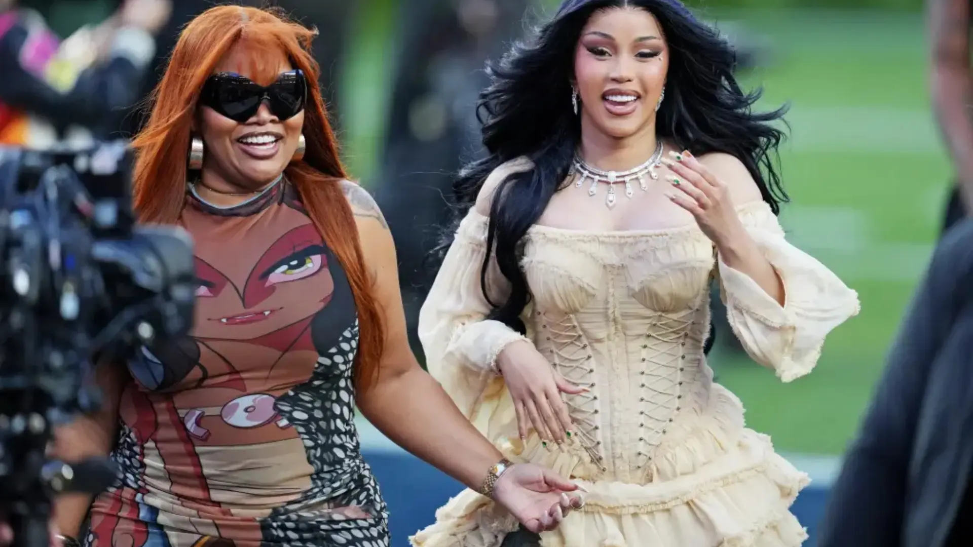 Cardi B walking on a field in a cream corset dress, holding hands with another woman near a camera crew.
