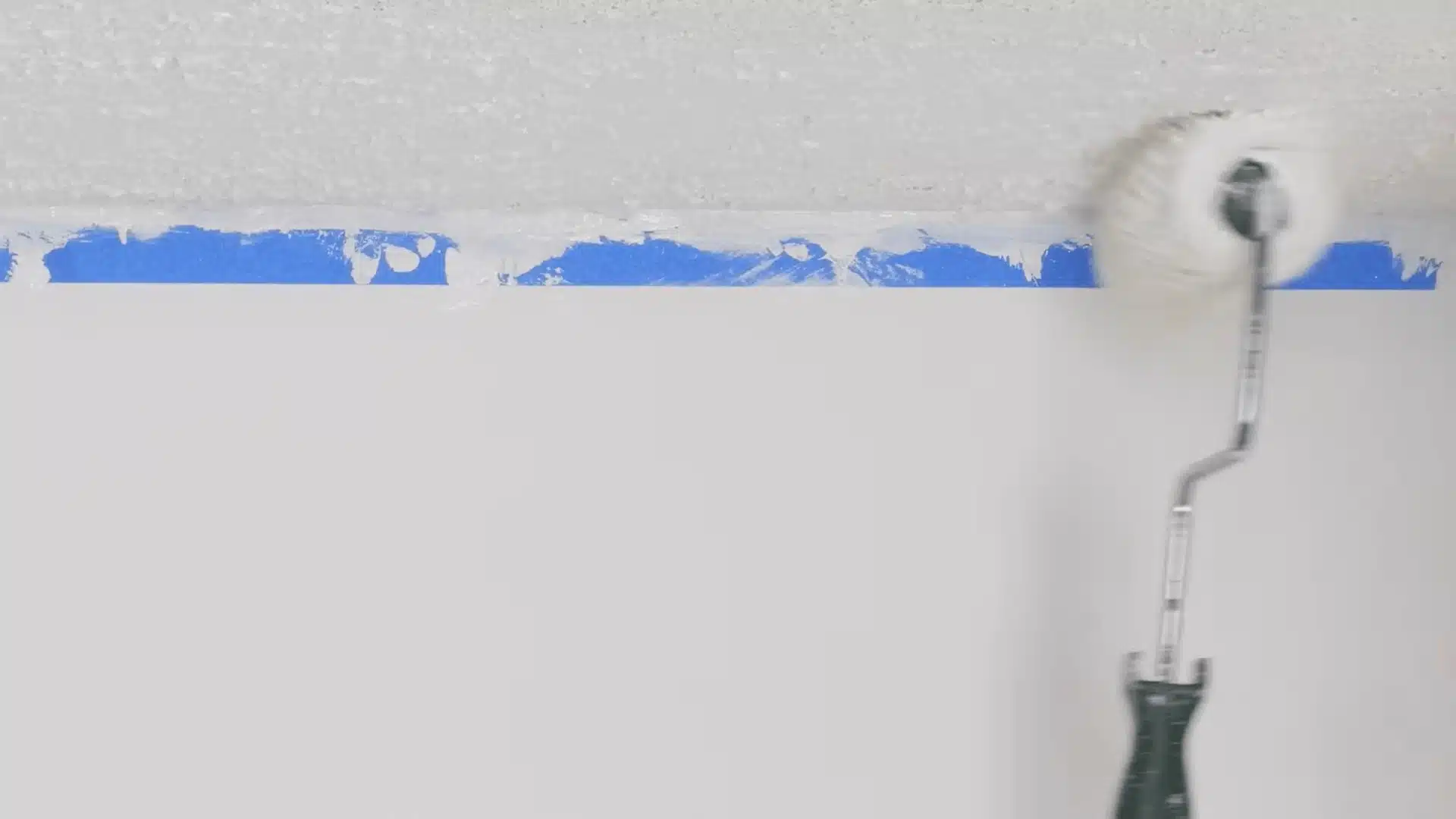 Paint roller applying a second coat to a popcorn ceiling near taped wall edges.