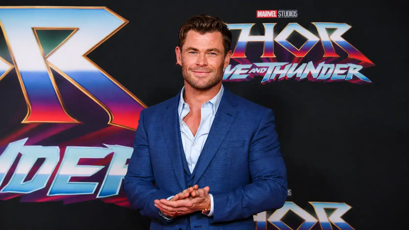 Chris hemsworth in a blue suit standing at a movie premiere event with a Thor themed backdrop behind him.