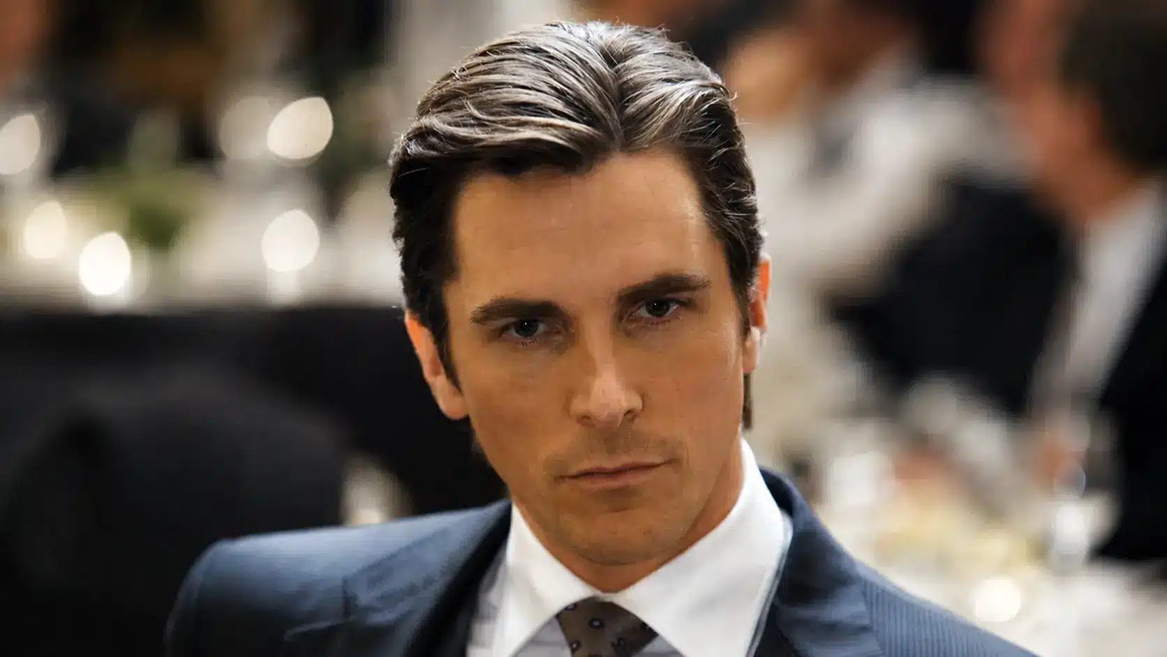 Christian bale in a dark suit and tie with slicked hair, seated in a dim restaurant with a blurred background.