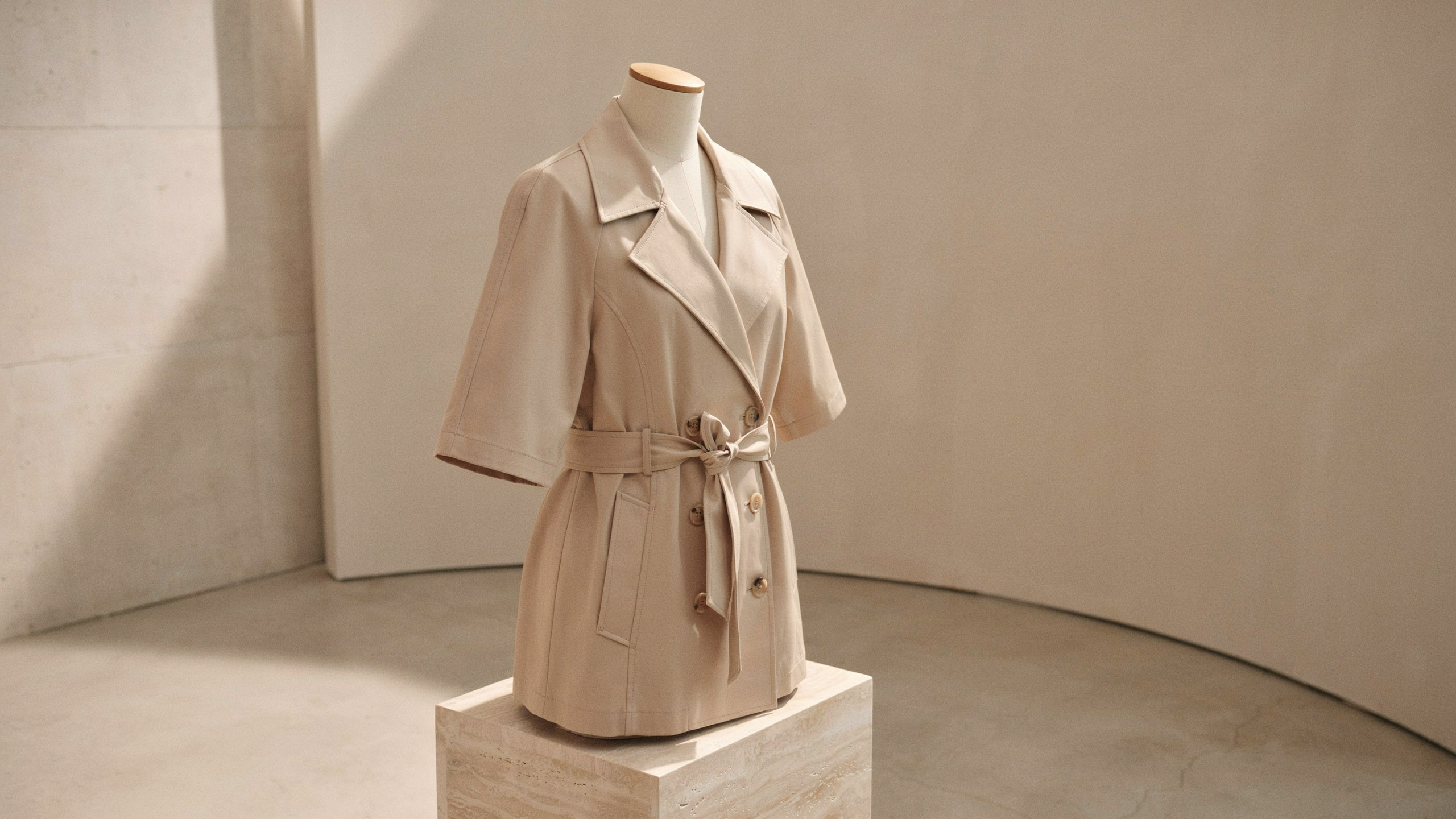 Cropped beige trench coat on a mannequin in a minimalist studio with soft natural light and curved walls.