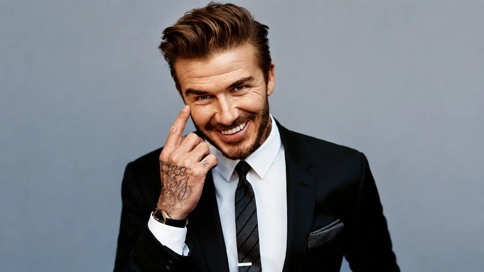 David beckham in black suit smiling and touching his face during a formal portrait photoshoot against a gray studio backdrop