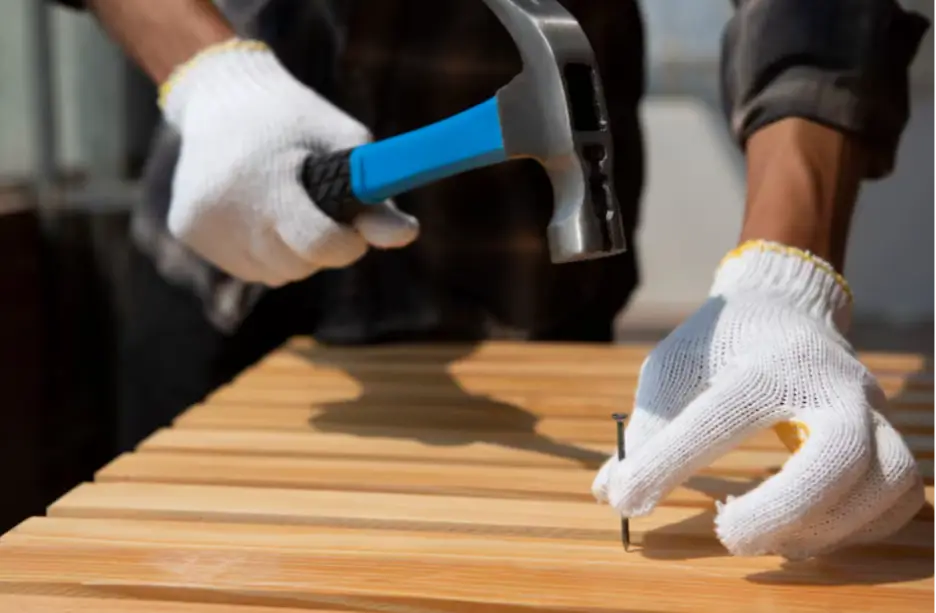 Worker wearing gloves hammering nail into wooden board outdoors