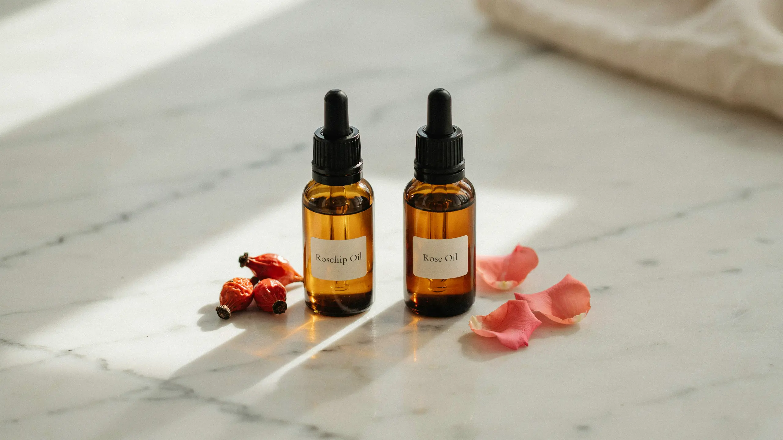 Amber rosehip oil bottles with droppers on marble surface with rose petals and dried rosehip fruits in soft light