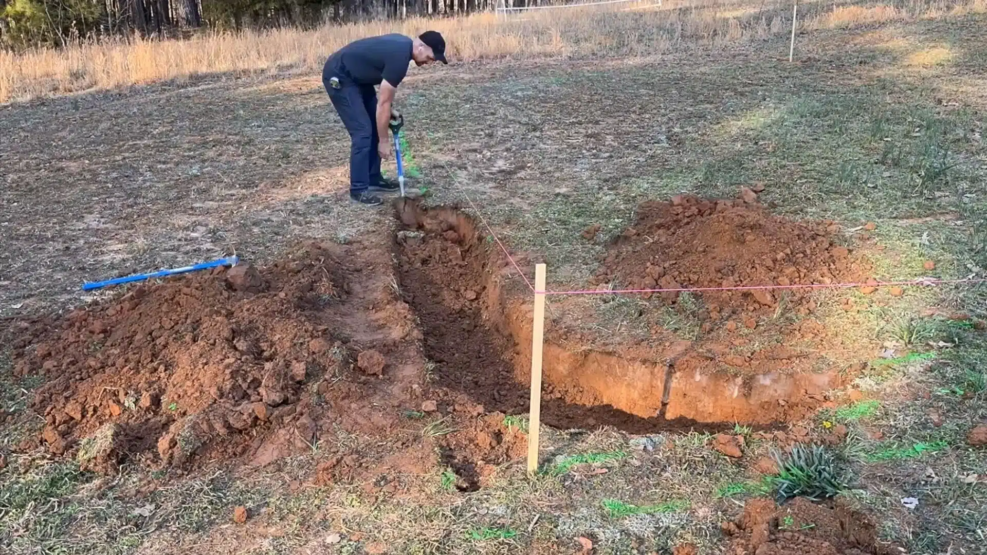 Person digging a trench in a marked garden area with stakes and string lines for layout.
