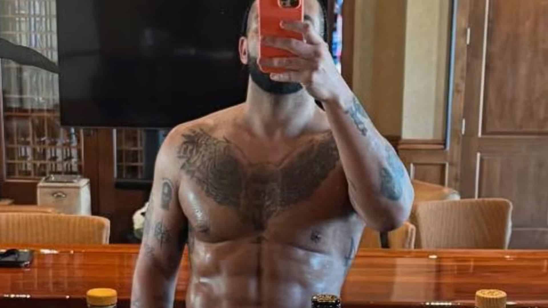 Shirtless Drake taking mirror selfie in gym holding phone over face showing defined abs and tattoos under lighting