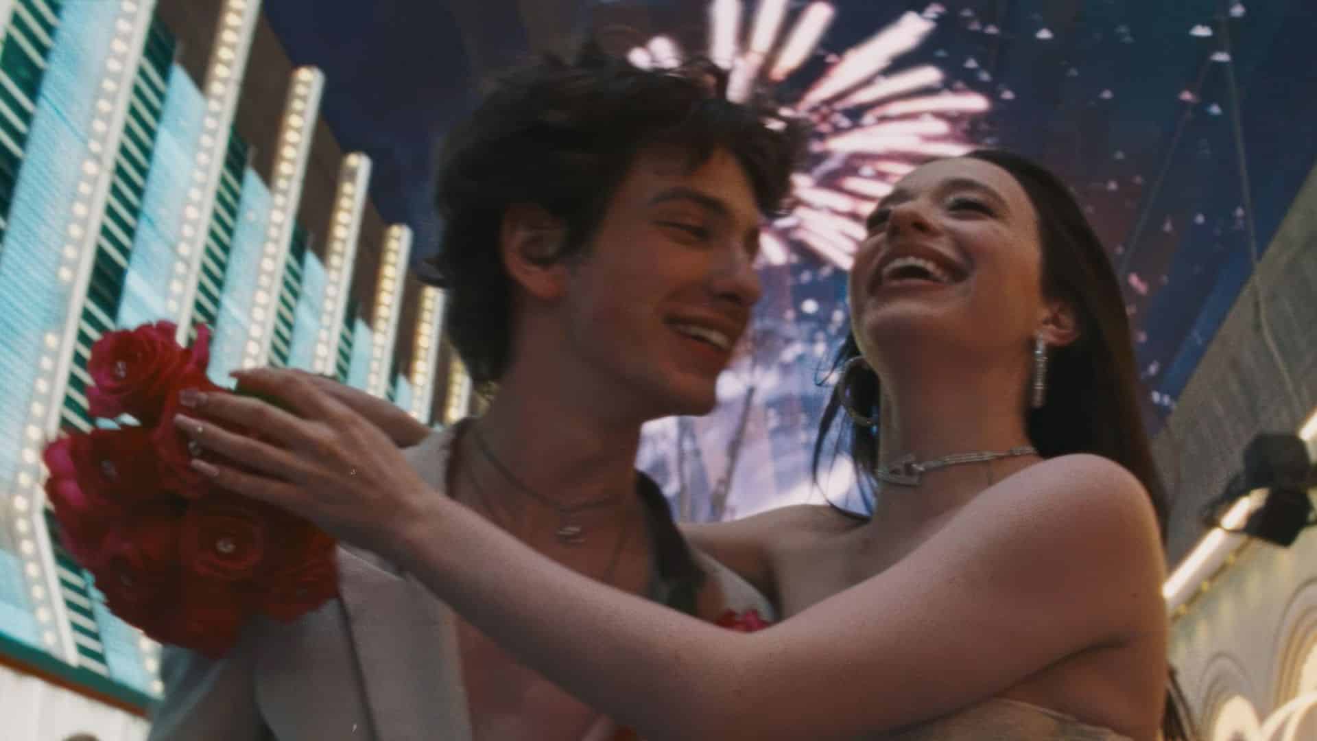 Young couple smiling and embracing on a neon-lit city street at night with fireworks overhead, holding roses