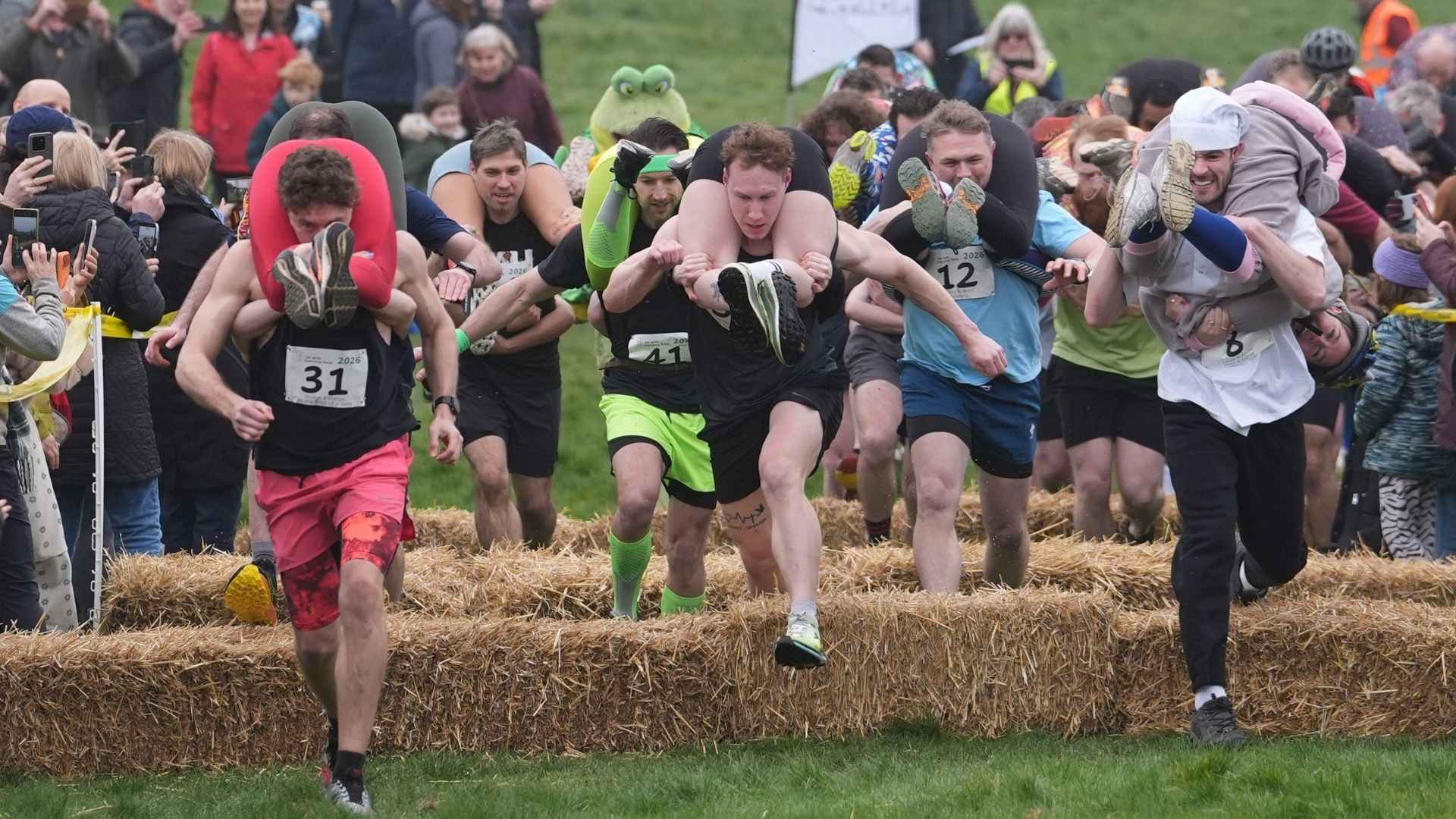 Runners carrying their wives on backs crossing hay bales in outdoor obstacle race crowd watching