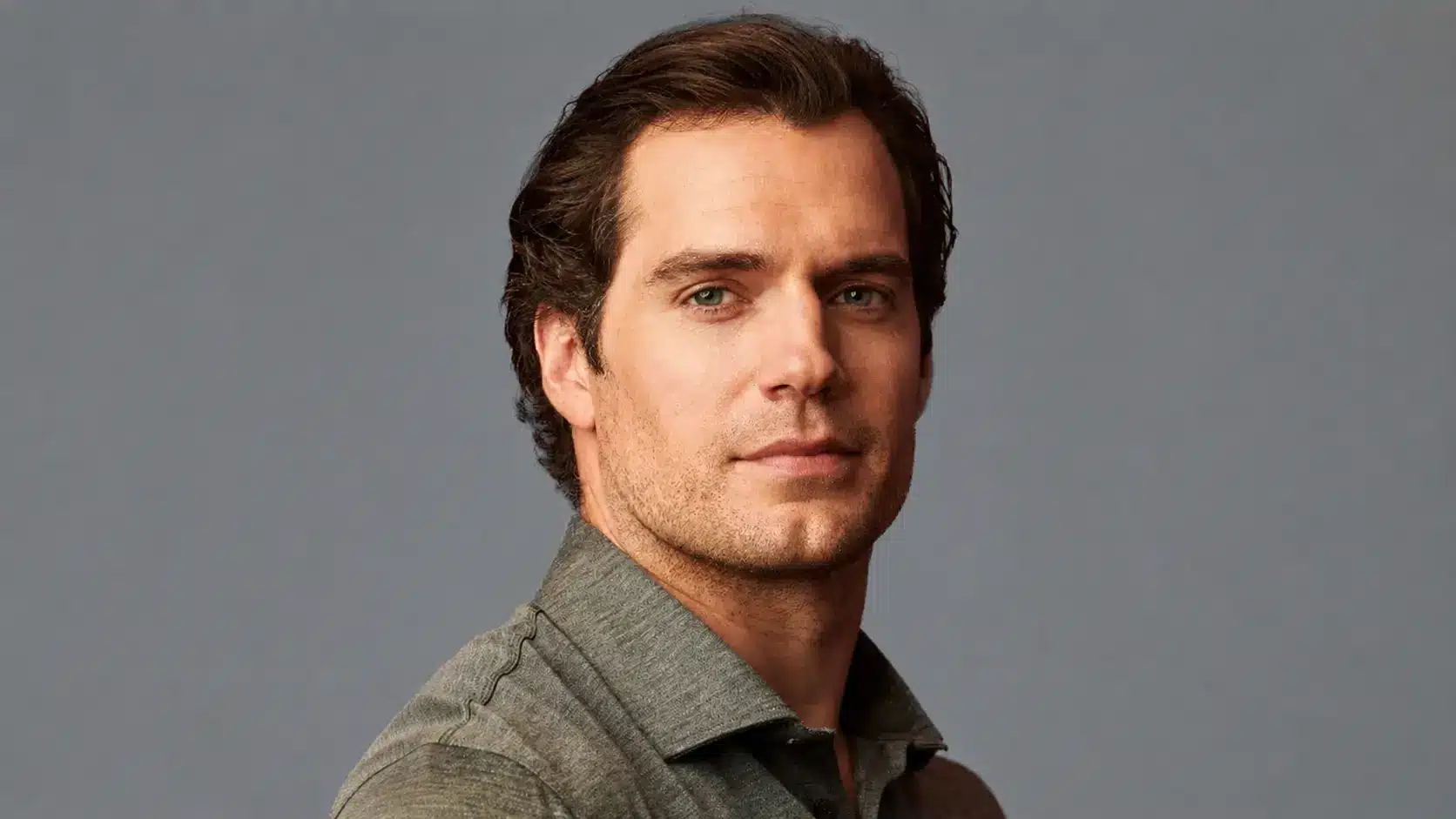 Portrait of henry cavill with dark hair and stubble wearing a gray shirt against a neutral gray background.