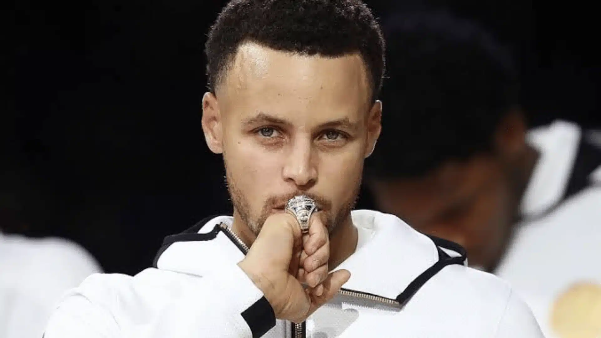 Stephen Curry wearing a white Warriors jacket while holding his championship ring.