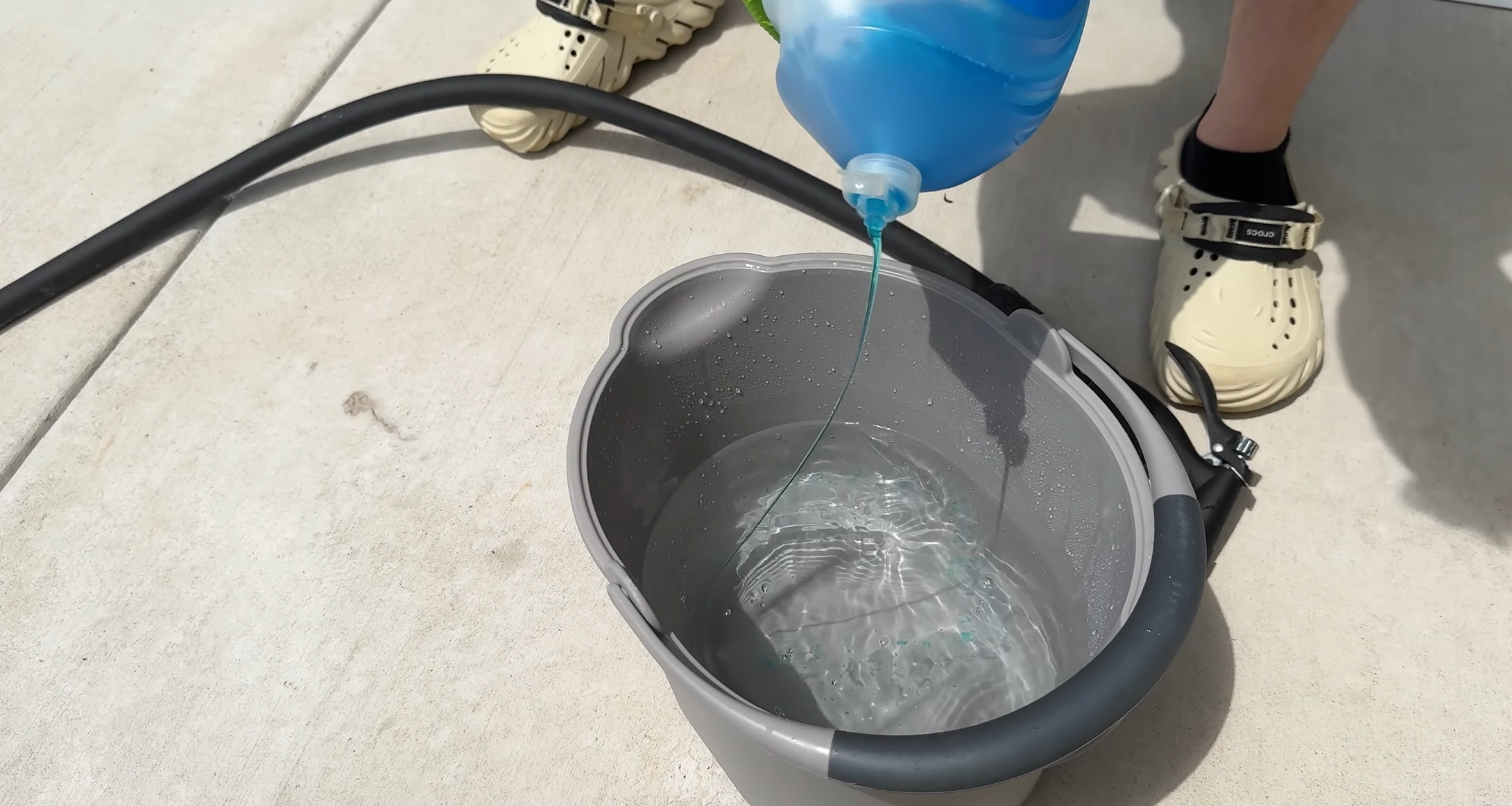 person pouring blue cleaning solution into a gray bucket of water on a sunlit patio