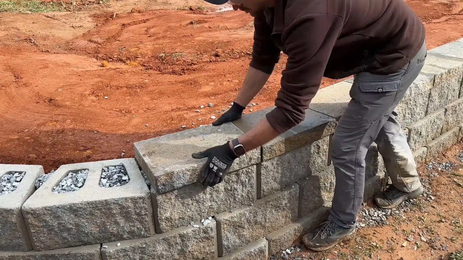 Person placing concrete blocks on a retaining wall during outdoor landscaping construction work.