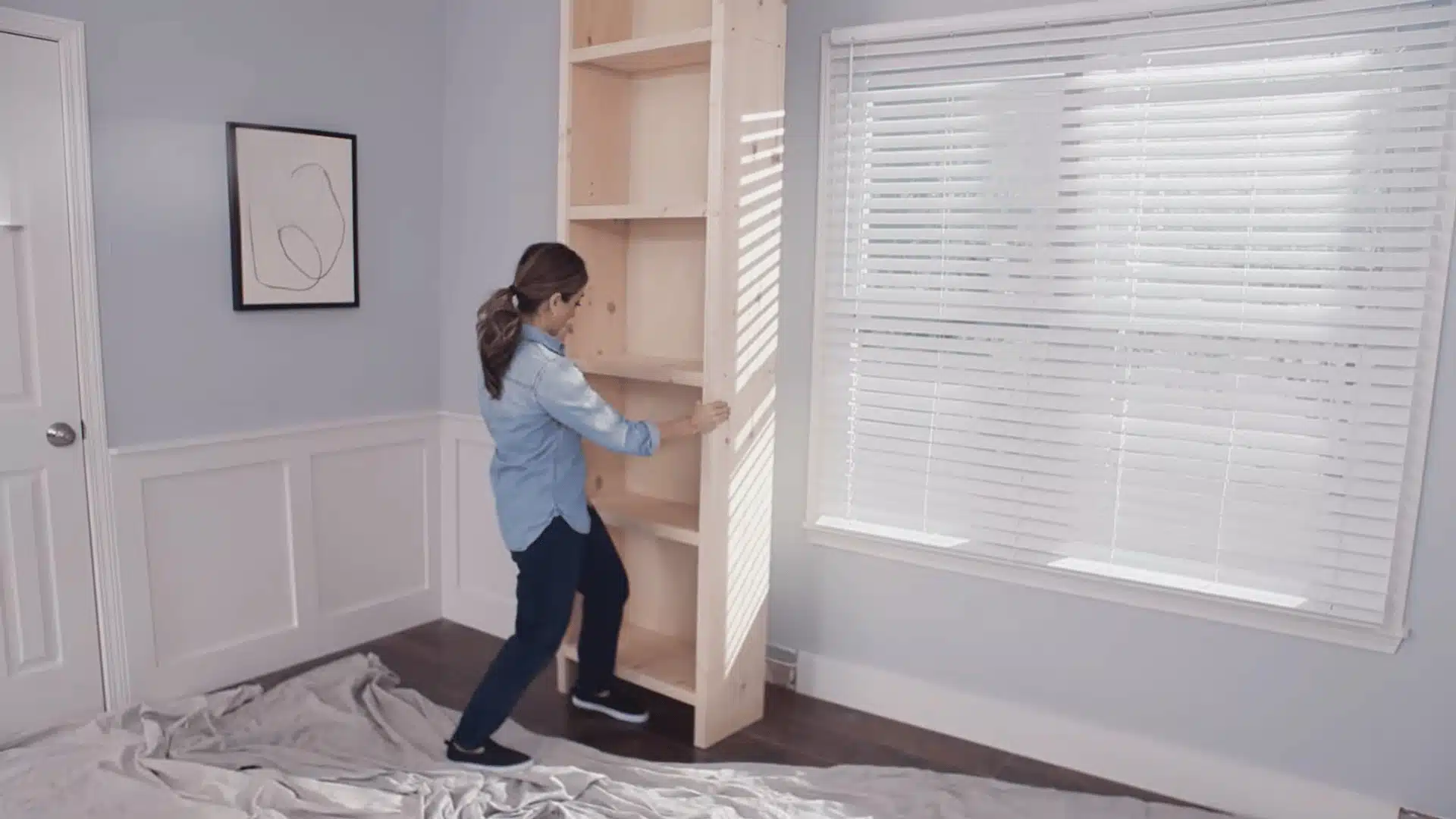 Person positioning a tall wooden shelving unit against a wall during a DIY installation project