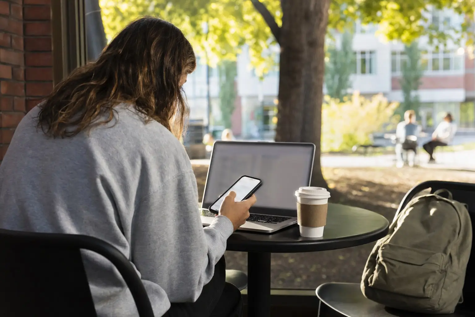 Long-haired person using smartphone and laptop at outdoor cafe table with coffee cup nearby