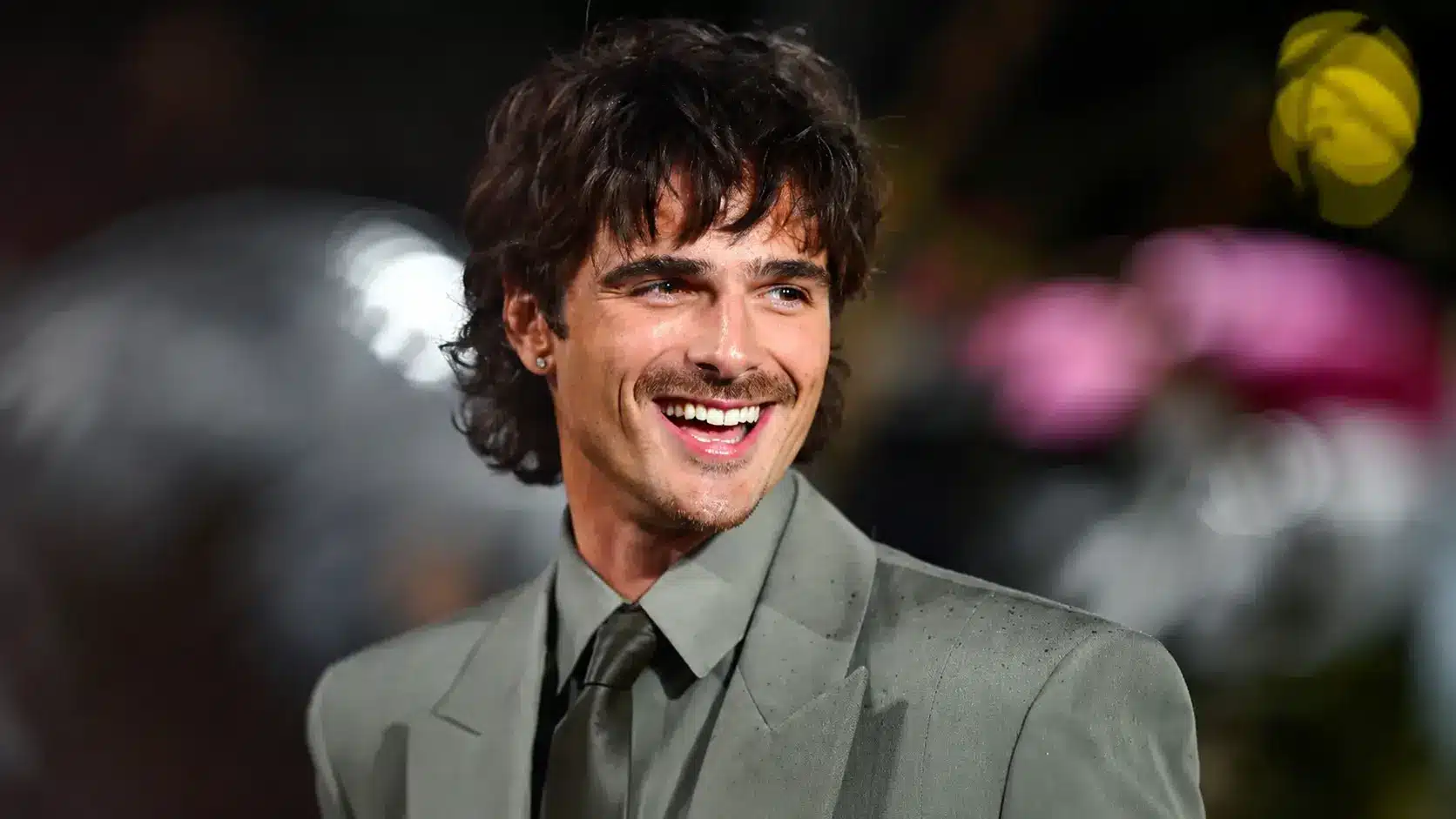 jacob elordi with wavy hair and mustache wearing a gray suit smiling at an event with a blurred background.