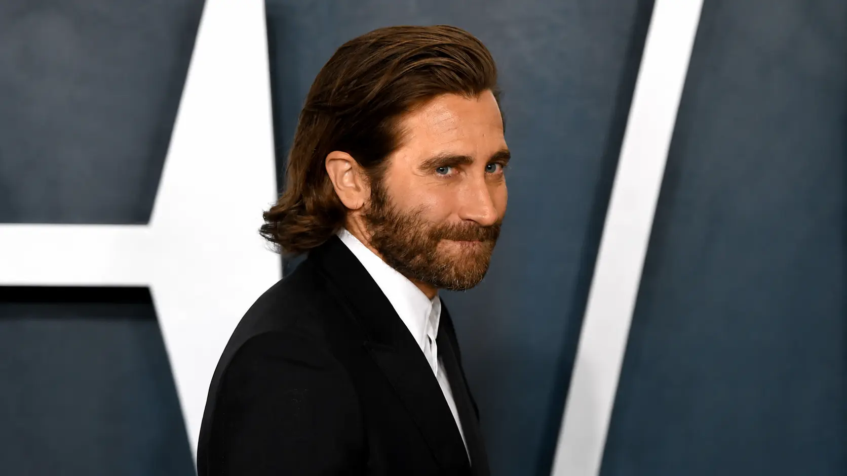 jake gyllenhall in a black suit with long hair and beard, posing at a formal event with a geometric backdrop.