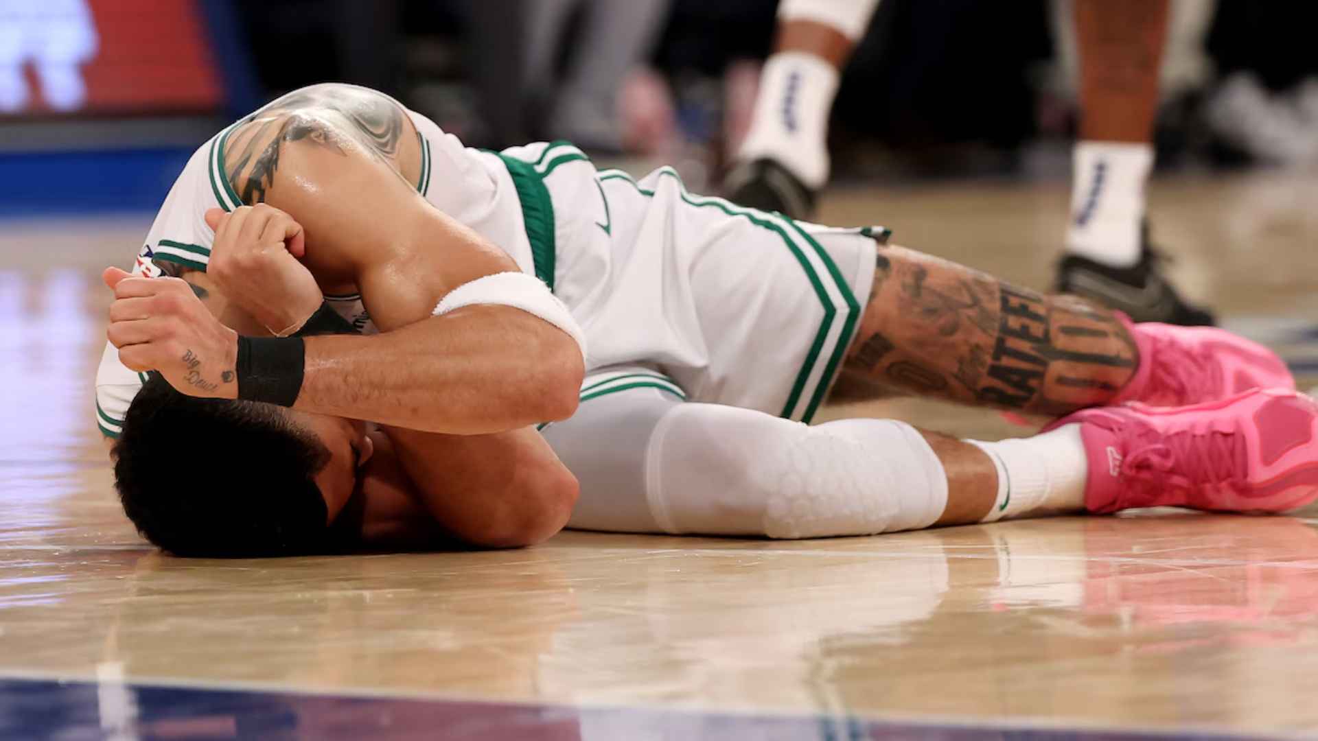Jayson Tatum lying on the basketball court in pain holding his leg during an NBA game in a Celtics uniform with pink shoes