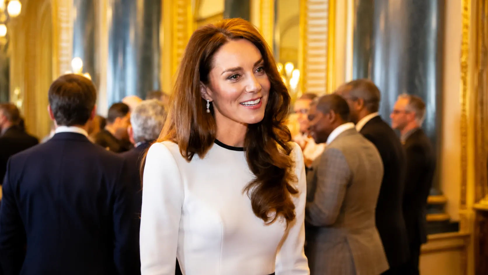 kate middleton in a white dress with black accents smiling at an indoor event with a blurred crowd background.