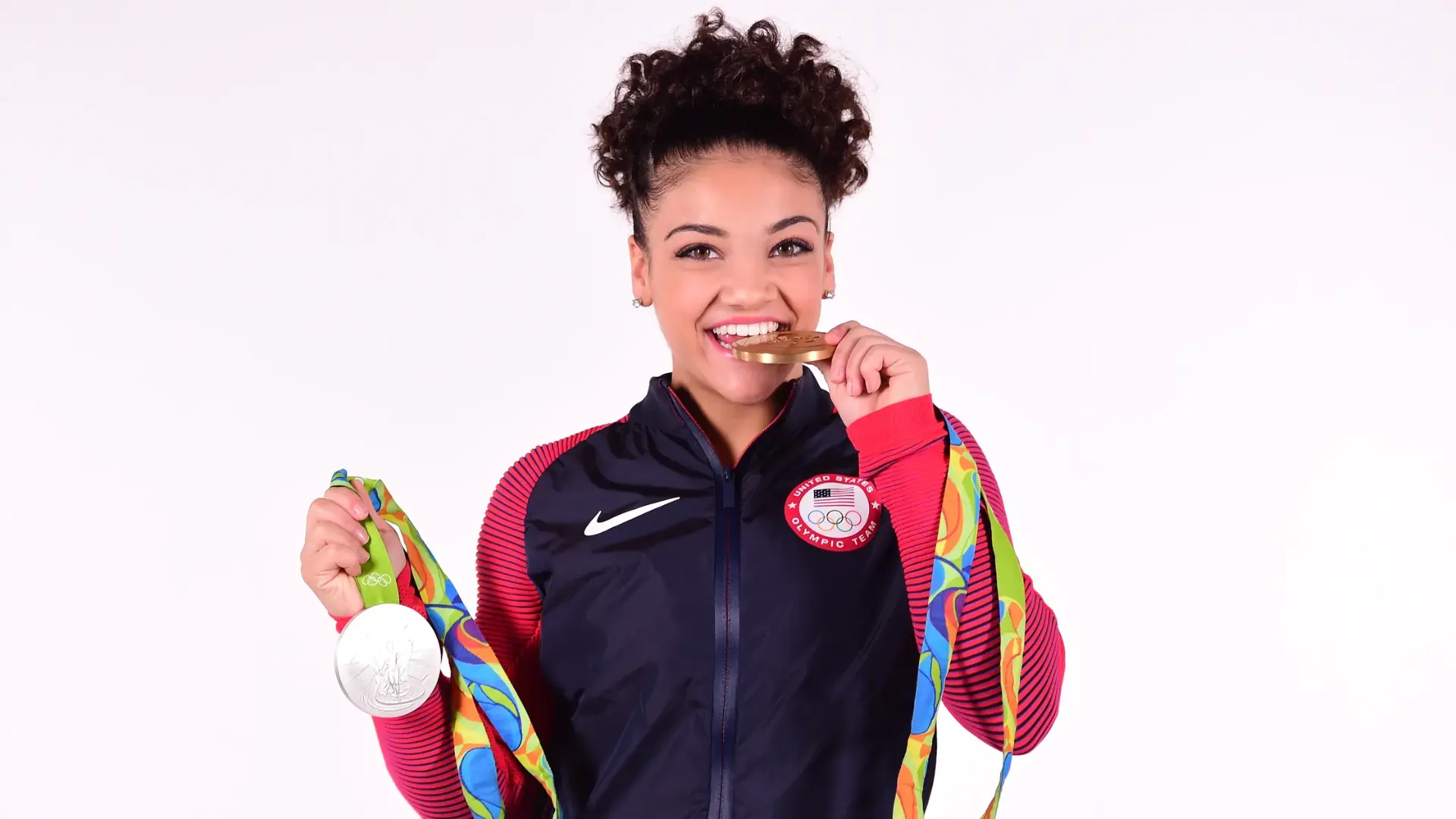 Smiling Olympic athlete holding two medals and biting a gold medal while wearing a Team USA jacket.