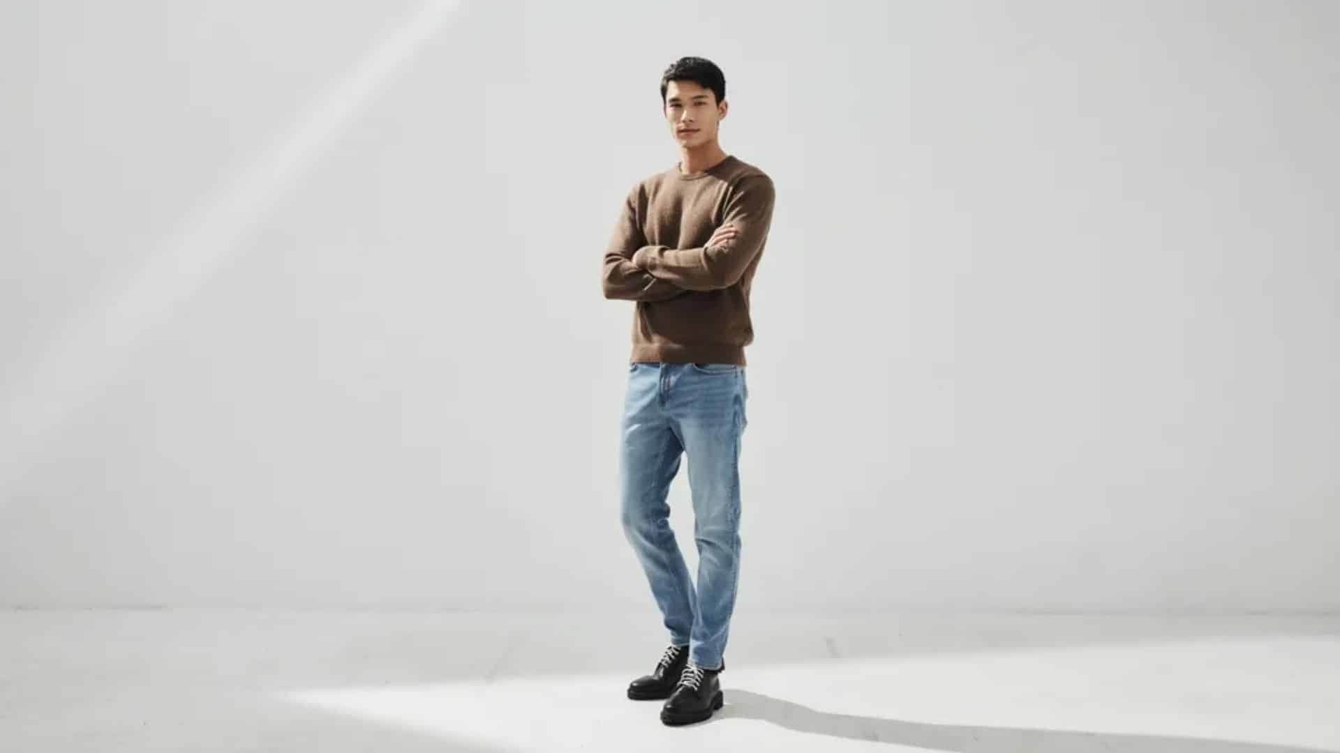 Man wearing brown knit sweater light blue straight jeans and black leather boots standing in minimalist studio pose