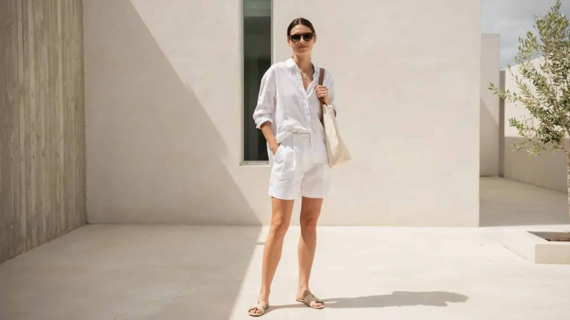 Woman wearing white linen shirt and shorts set with flat sandals and tote bag standing in minimal sunny courtyard scene