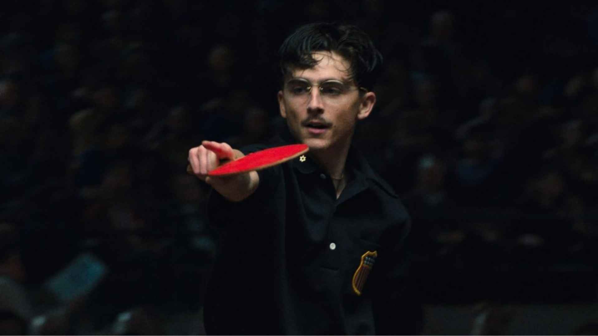 Timothee Chalamet in glasses playing table tennis, extending a red paddle forward during a focused match.