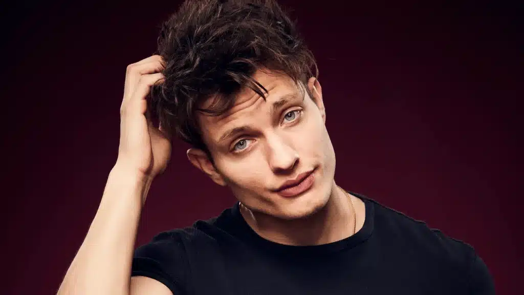 Portrait of a man with dark hair wearing a black shirt, posing against a dark background in a studio setting.