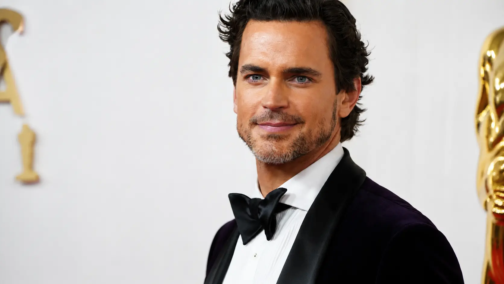 Close up red carpet portrait of Matt bomer in black tuxedo and bow tie smiling against awards backdrop with soft lighting