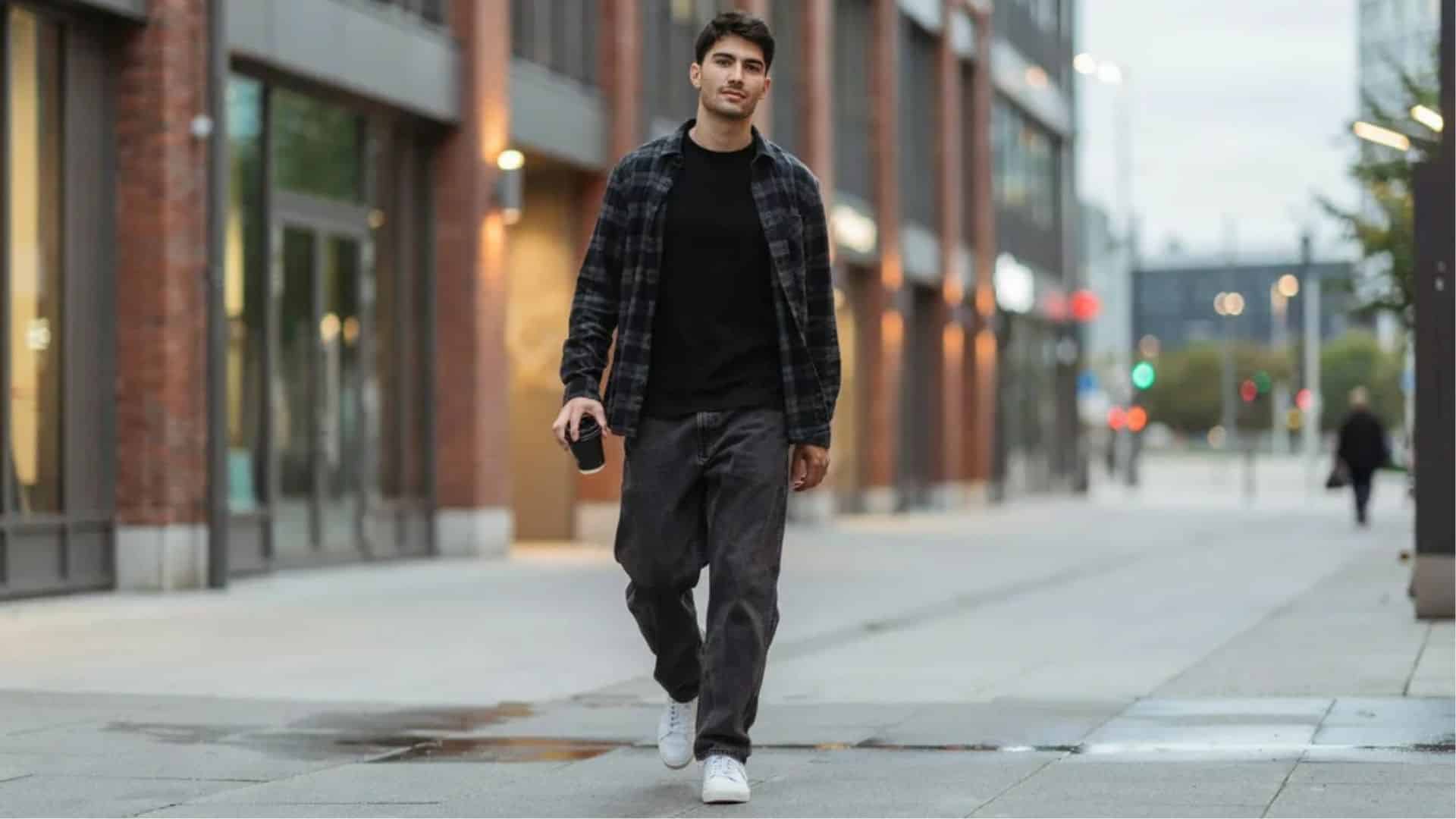 Man with visible face wearing plaid overshirt black tee baggy dark jeans and white sneakers walking city street with coffee cup.