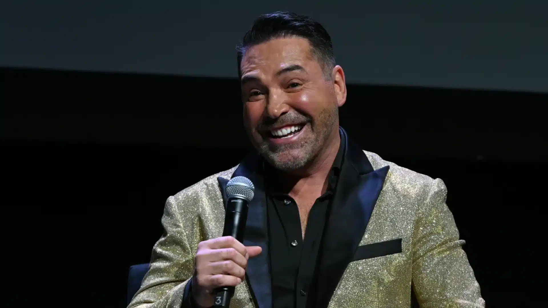 Man speaking into a microphone while wearing a sparkling jacket at a public event.