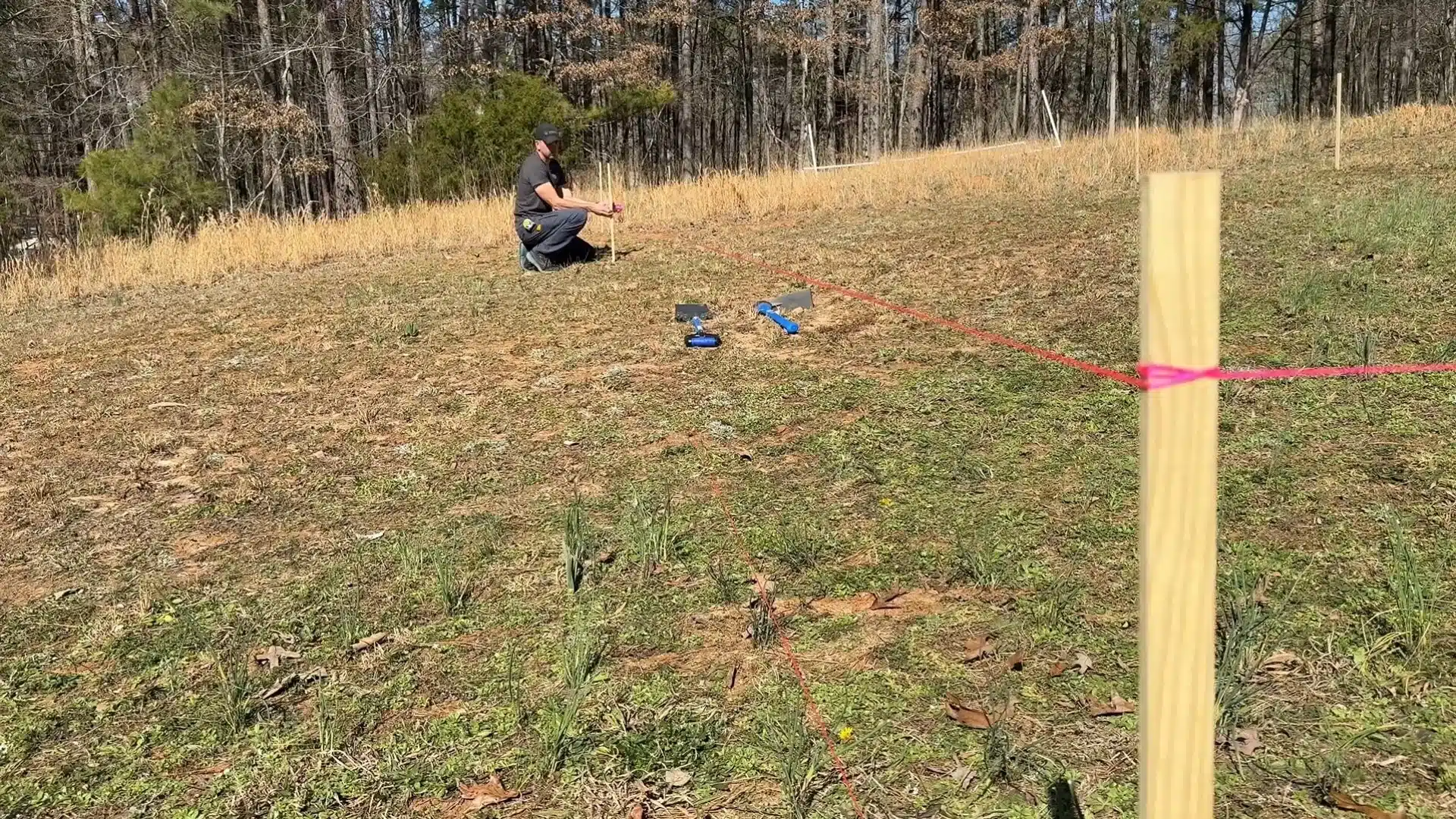Man measuring a marked garden plot with stakes and string on a grassy hillside.