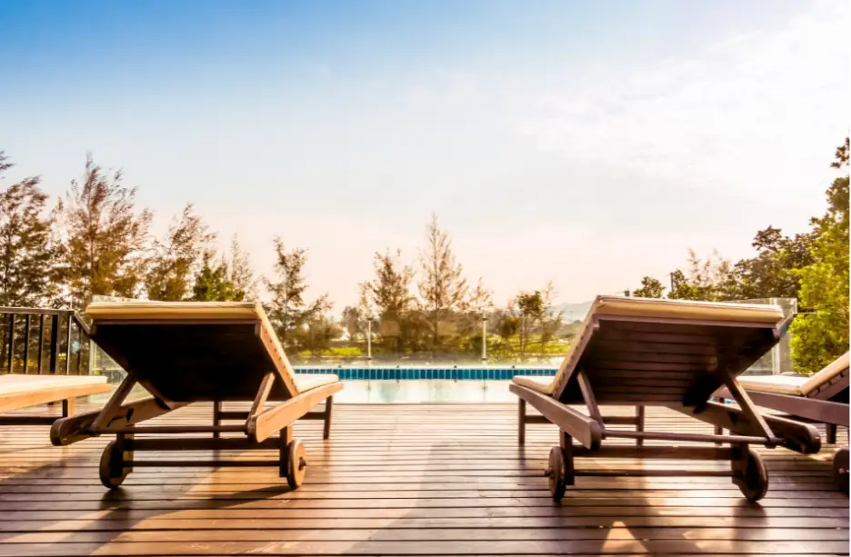 Wooden deck chairs facing pool in sunny outdoor setting with trees