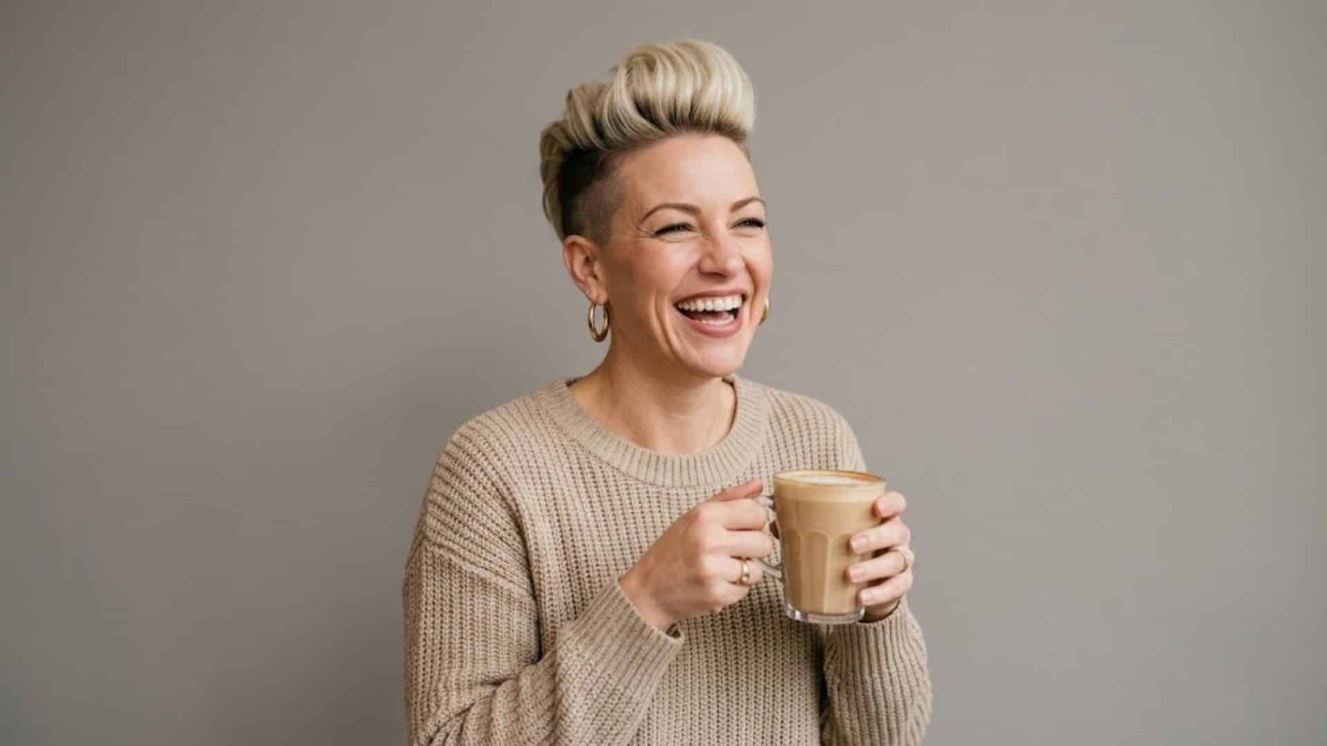 Woman with a blonde quiff haircut and shaved sides, laughing while holding a coffee mug