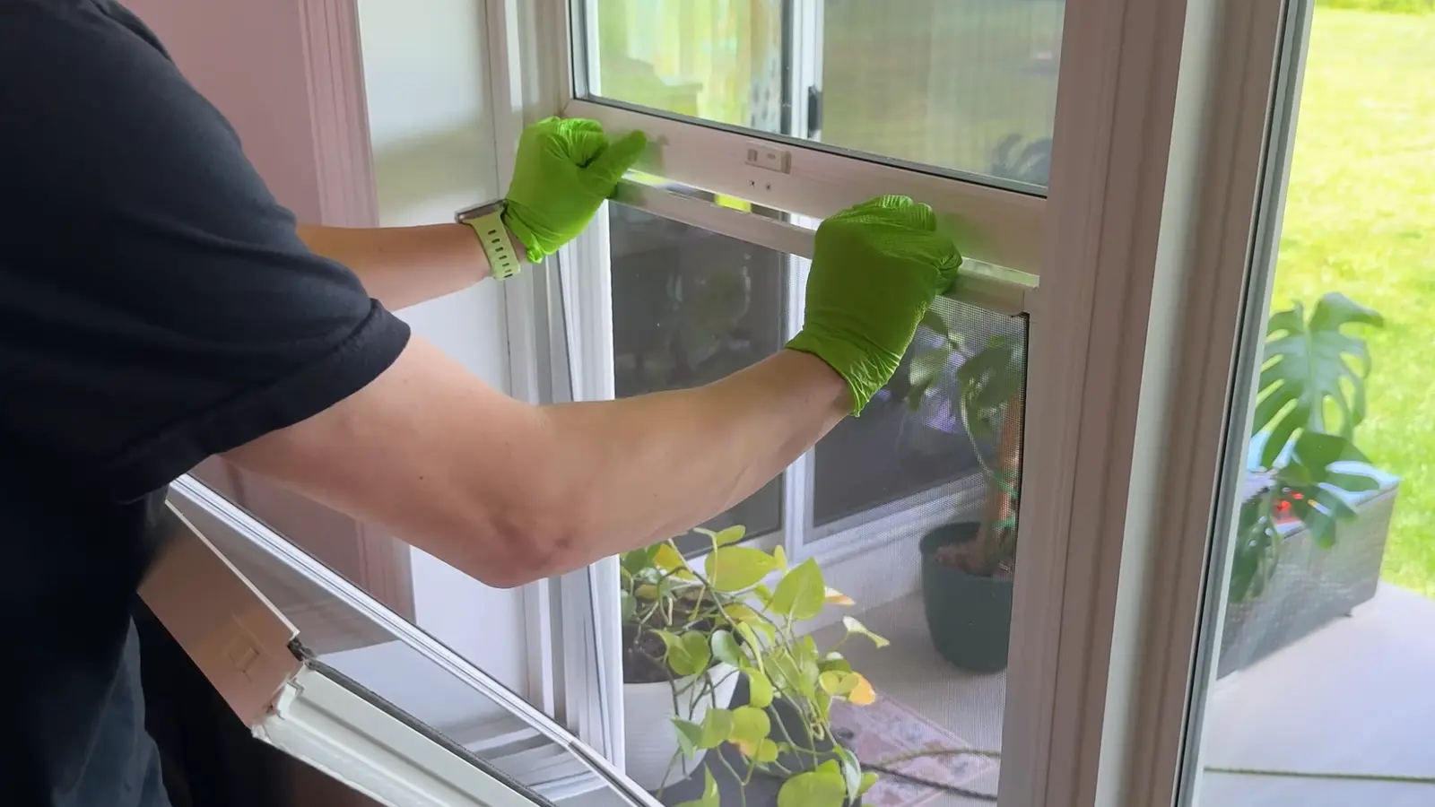 gloved hands gripping both sides of a white window screen, tilting it inward to remove it from the frame