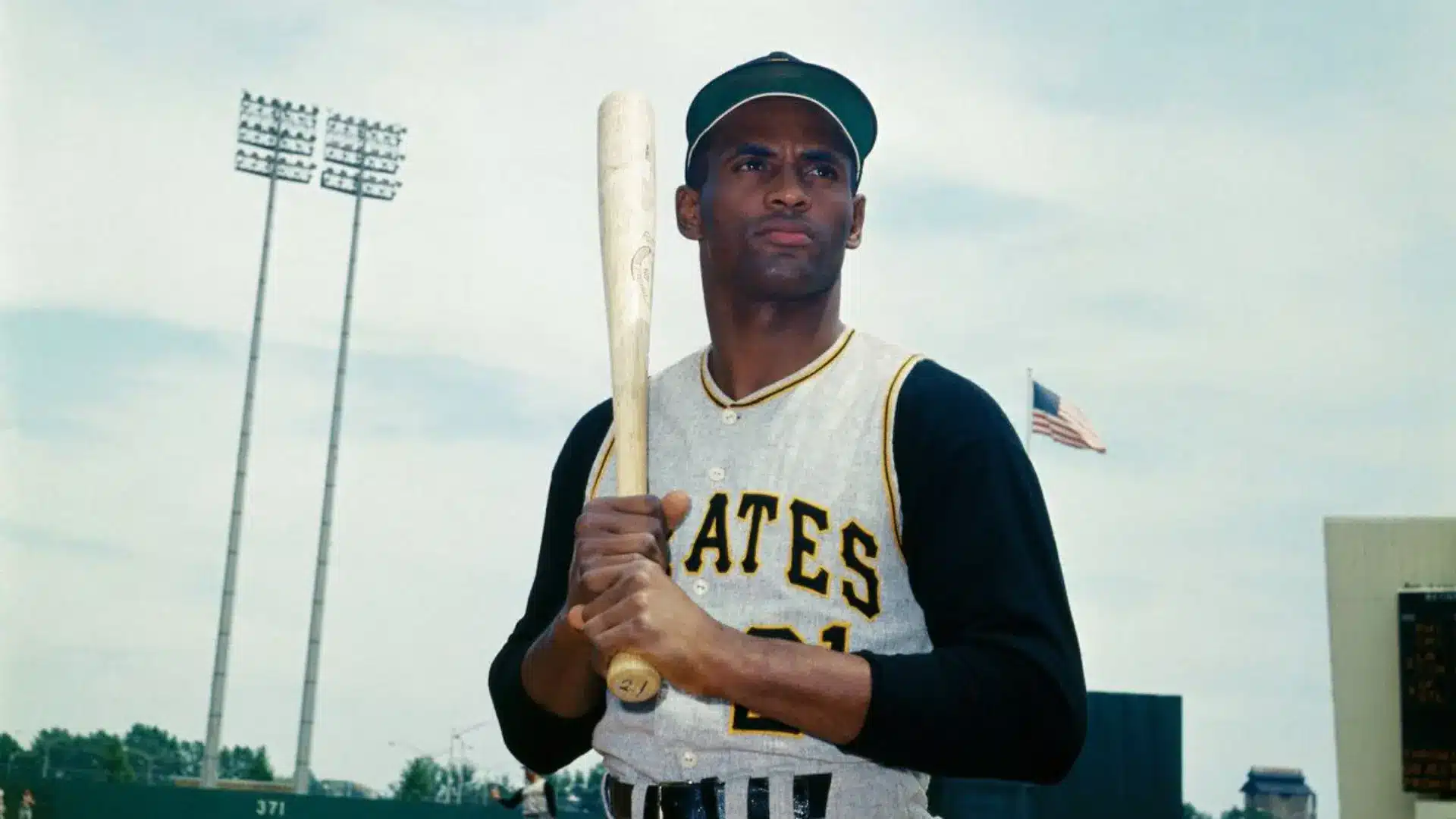 Baseball player holding a bat while wearing a vintage uniform on a stadium field.