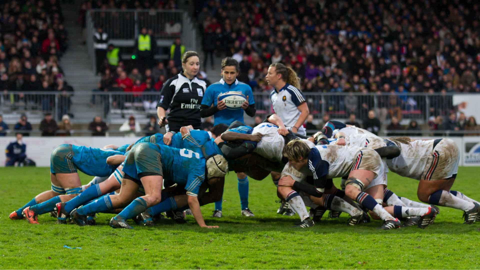 Women's rugby teams engage in a powerful scrum during a competitive match as the referee watches closely.