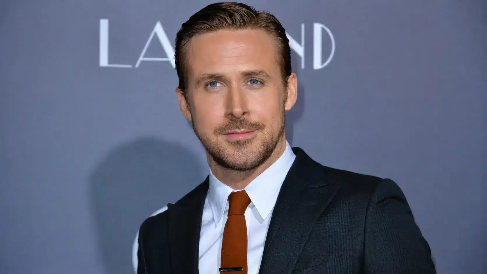 Close up portrait of ryan Gosling in dark suit white shirt and brown tie posing at red carpet backdrop with LA LAND text shot