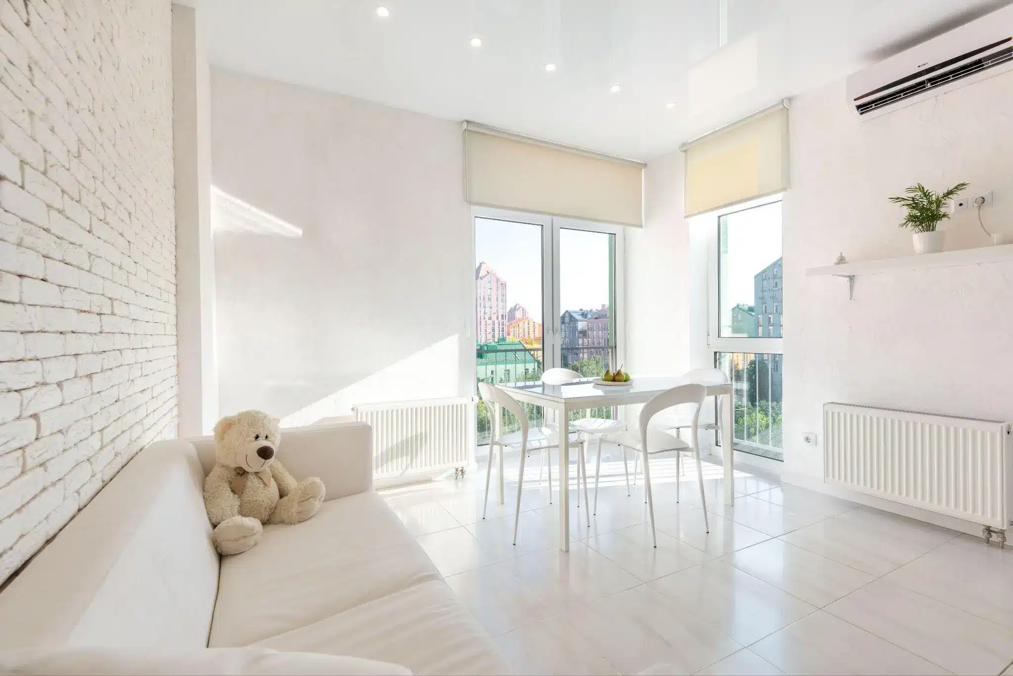 Bright, modern living room with white sofa, teddy bear, and dining table by large windows