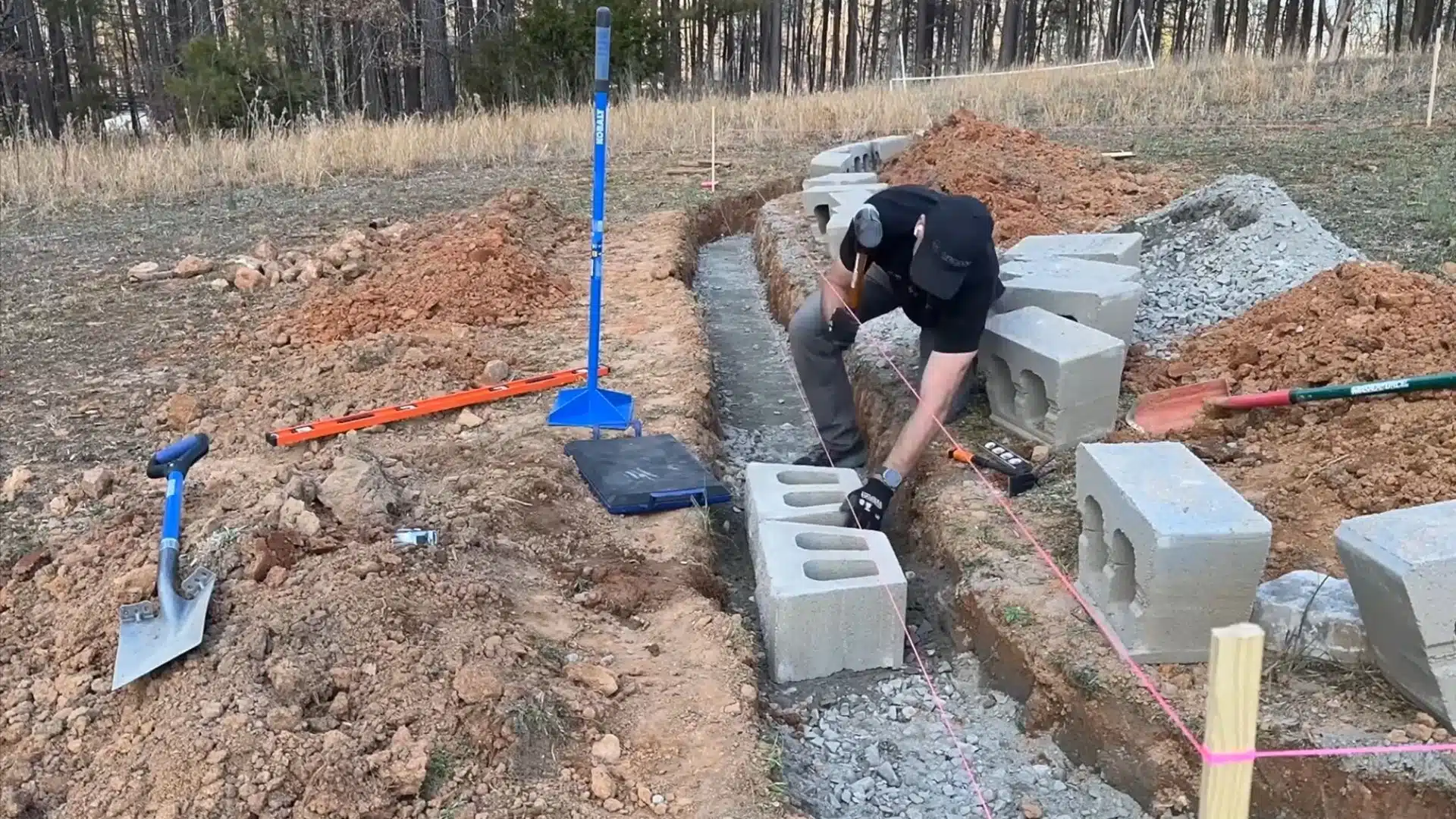 Person installing concrete blocks in a dug trench with gravel base, tools, and layout strings at a construction site.