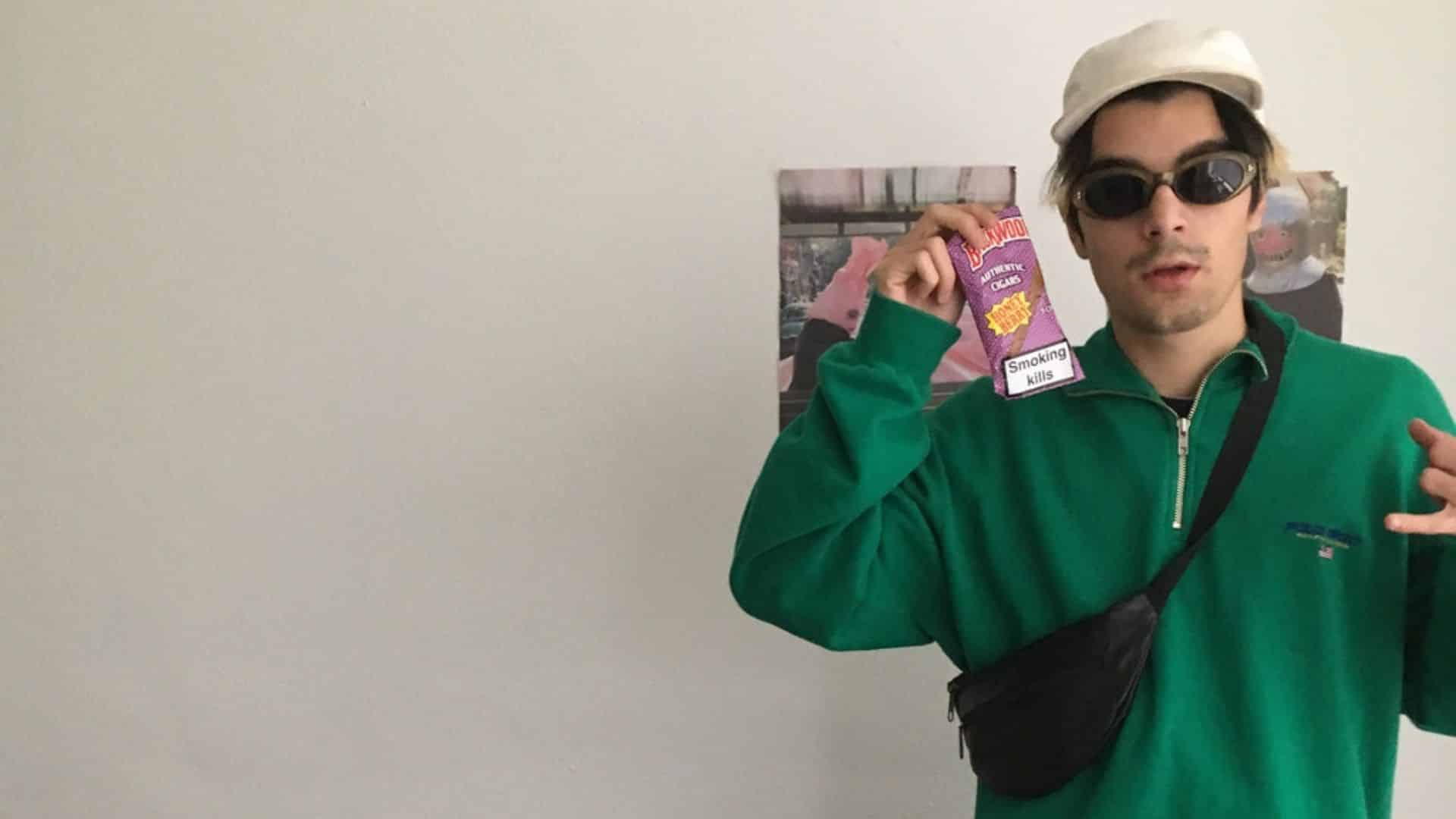 Young man in green sweatshirt, sunglasses and cap holding cigarette pack labeled Smoking Kills against plain wall indoor
