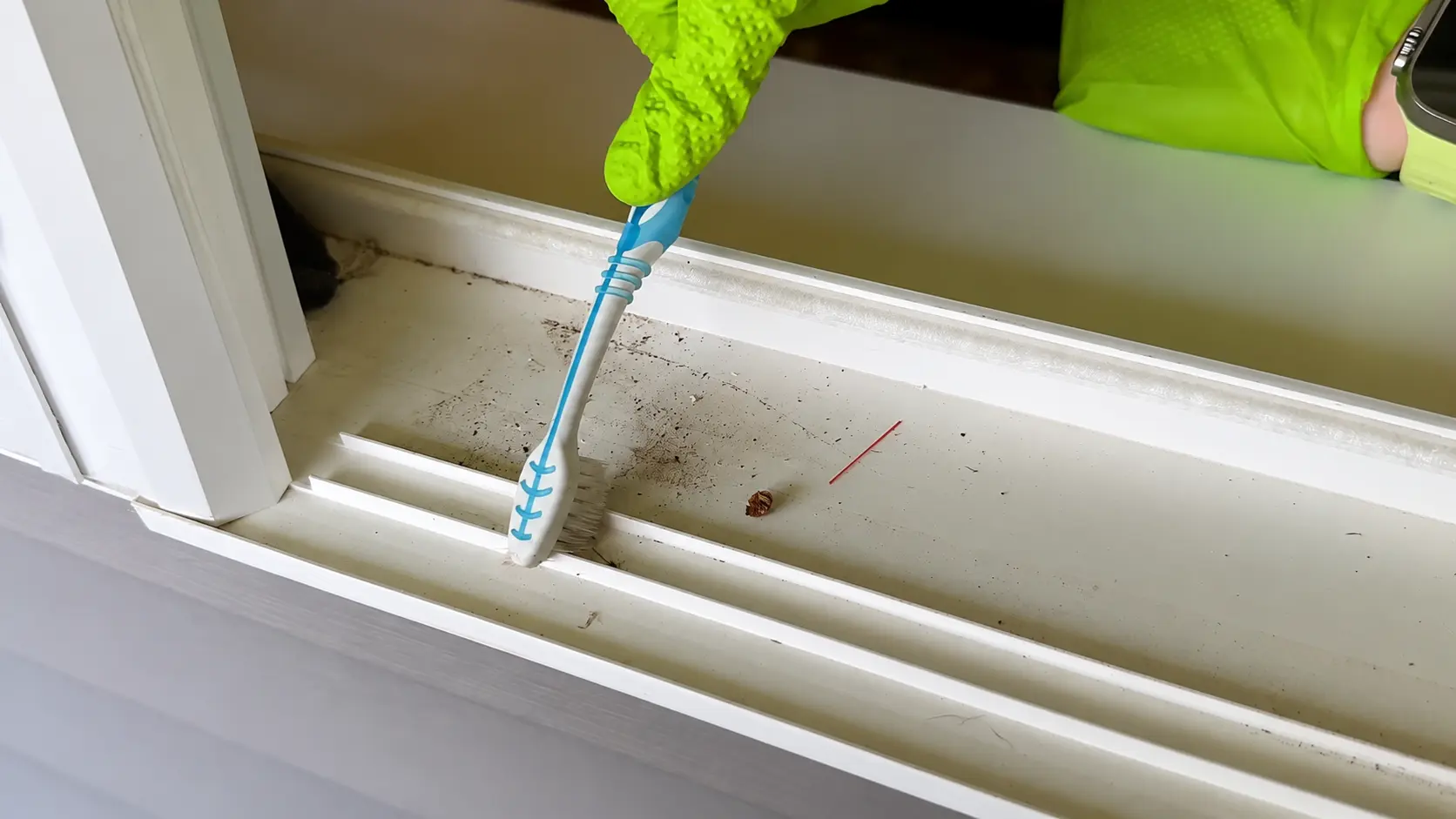 gloved hand scrubbing dirt from a white window track with a blue and white toothbrush