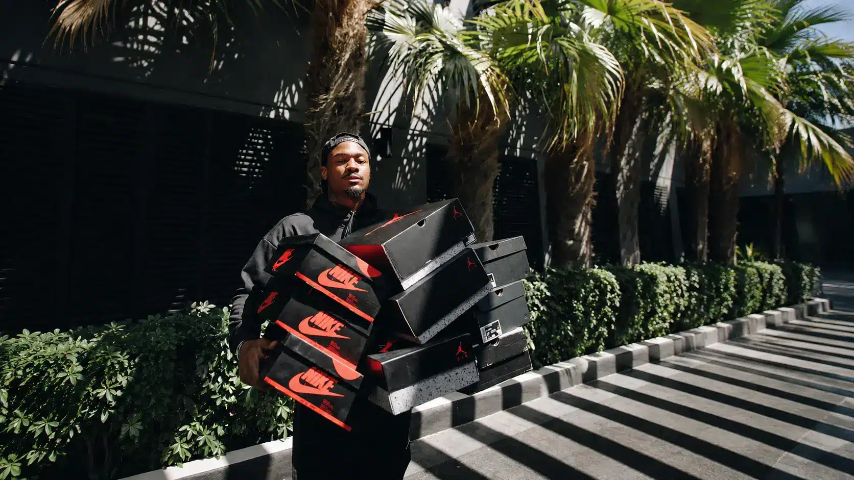 Stefon Diggs carrying stacked Nike shoe boxes outdoors near palm trees on striped walkway in bright sunlight scene.
