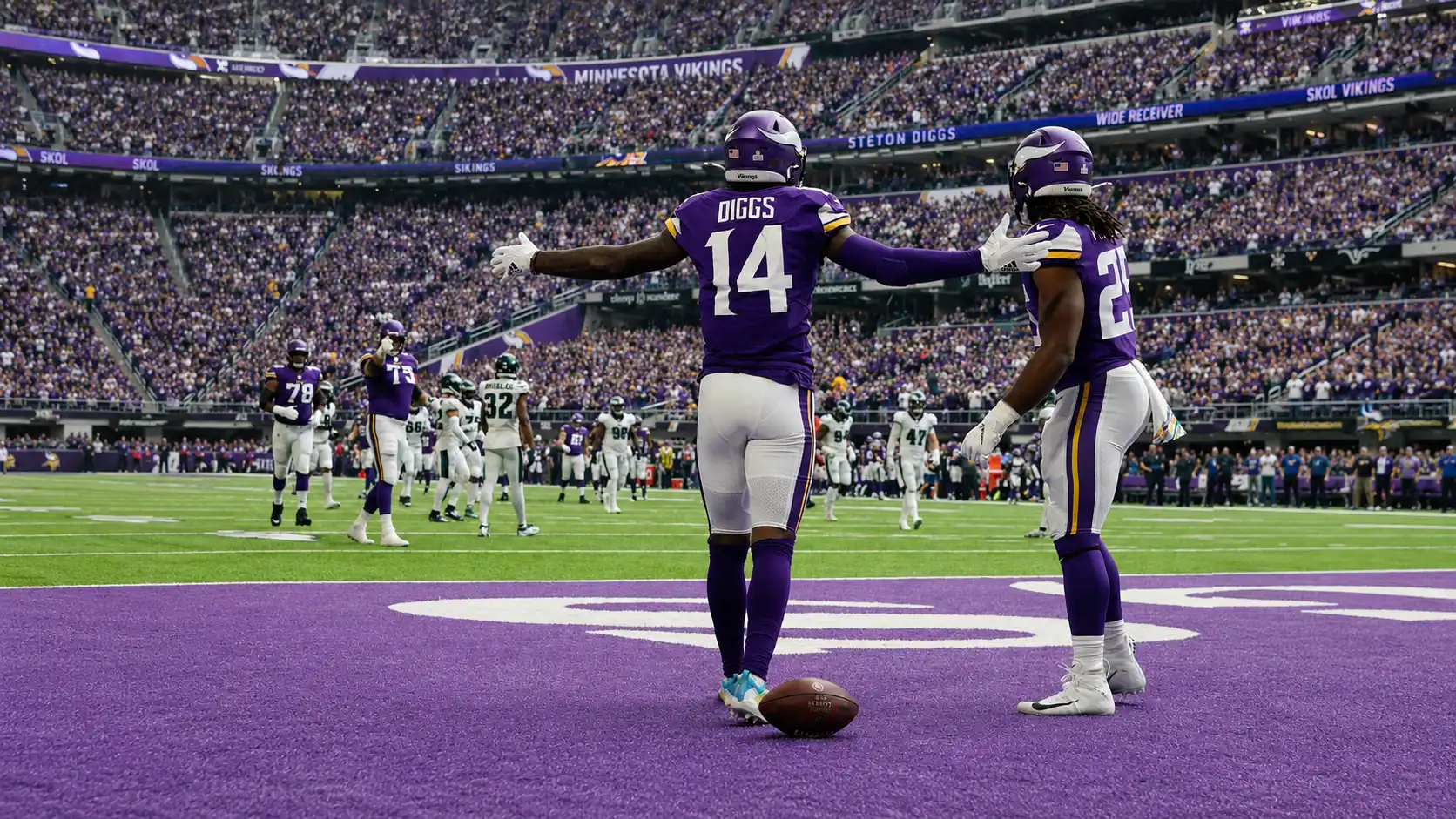 Two football players in purple uniforms standing on the field during a game inside a packed stadium.