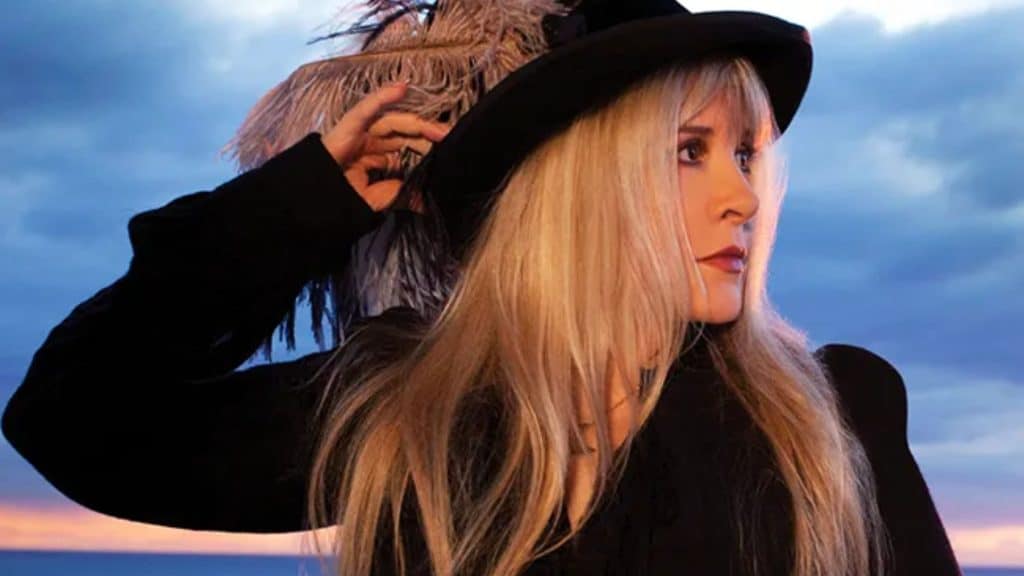 Stevie Nicks outfits inspired look featuring a black wide brim hat, dark layered clothing, and long blonde hair against a sunset sky background