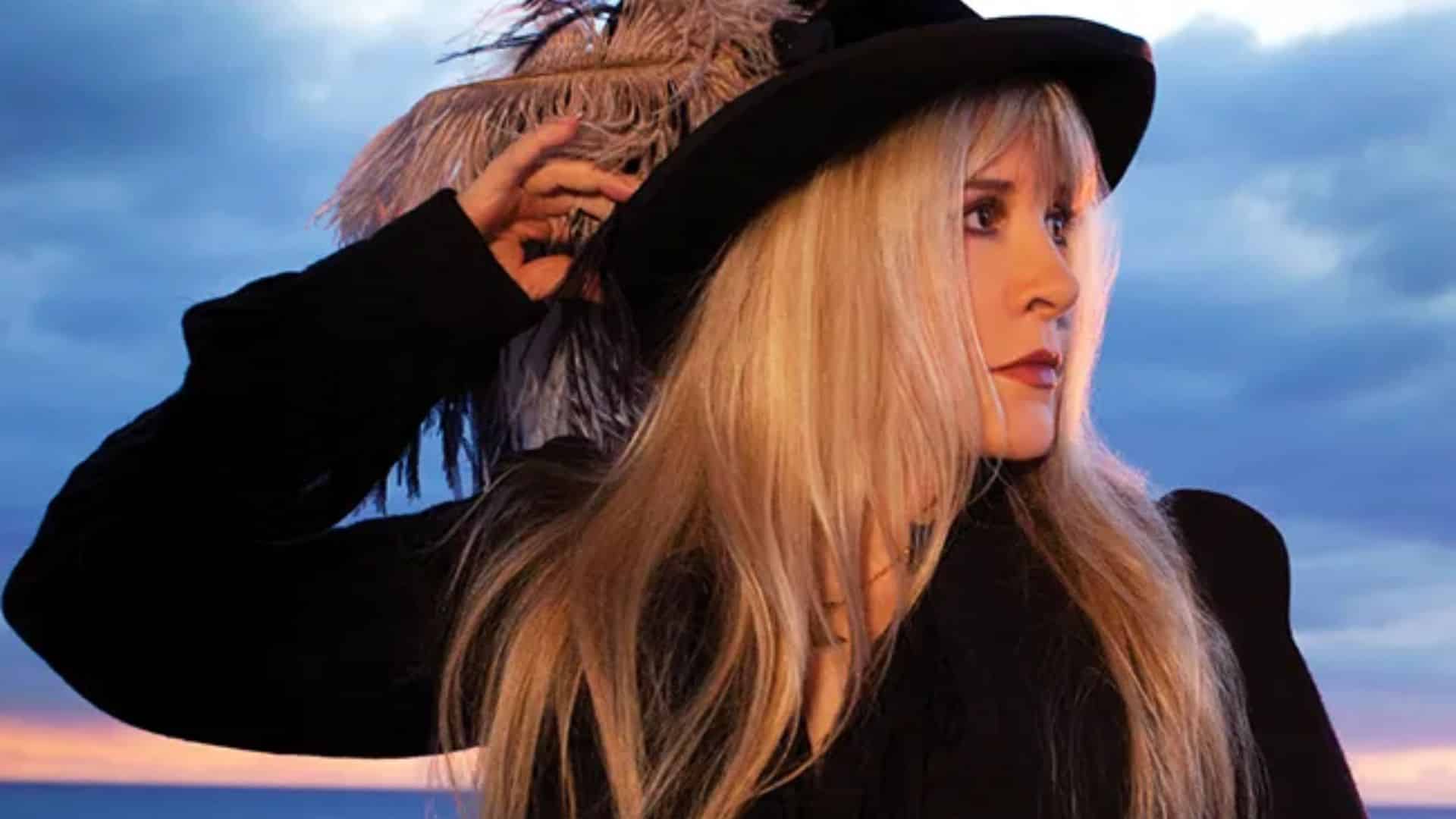 Stevie Nicks outfits inspired look featuring a black wide brim hat, dark layered clothing, and long blonde hair against a sunset sky background