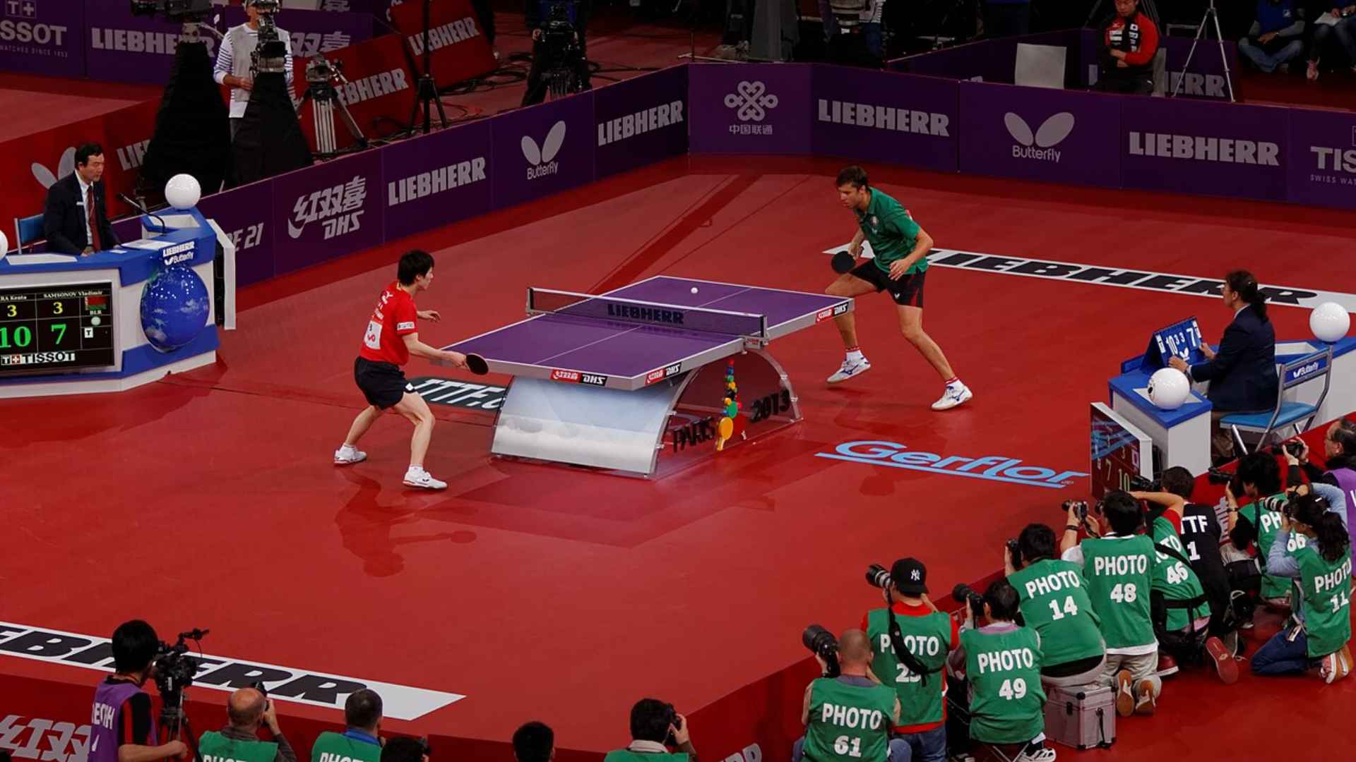 Two table tennis players compete in a fast-paced international match as officials, photographers, and fans look on.