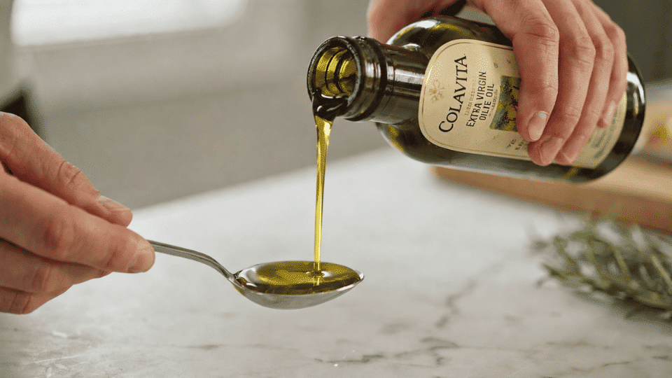 Close up of olive oil being poured from a branded bottle into a tablespoon with hands holding both items in a kitchen setting