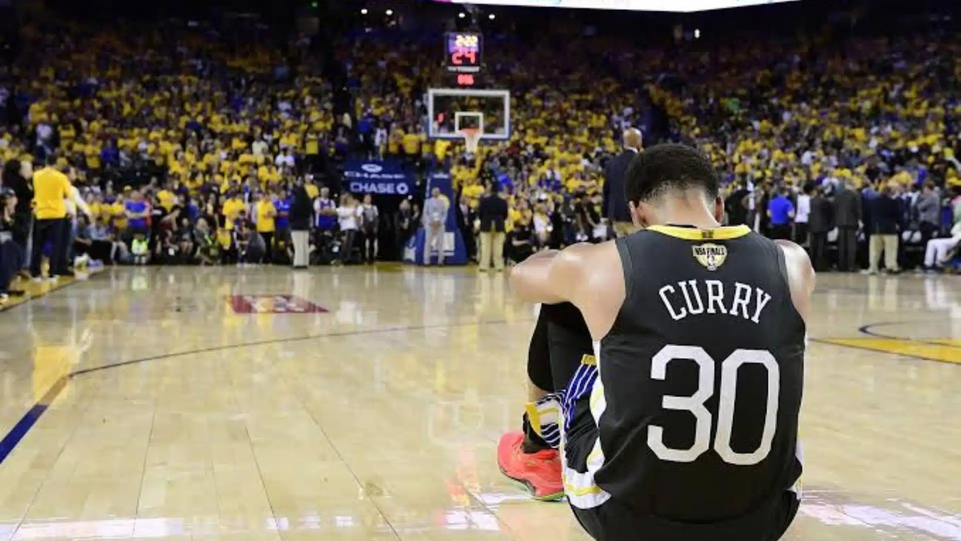 Basketball player sitting on the court in a black jersey during a game with a crowd in the background.